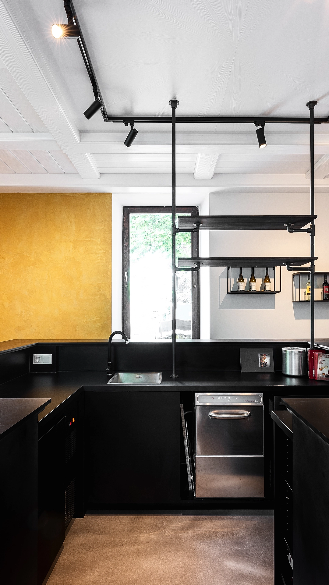 Modern kitchen with black countertops and cabinets, a stainless steel dishwasher, a small sink under a window, and a mustard yellow accent wall.