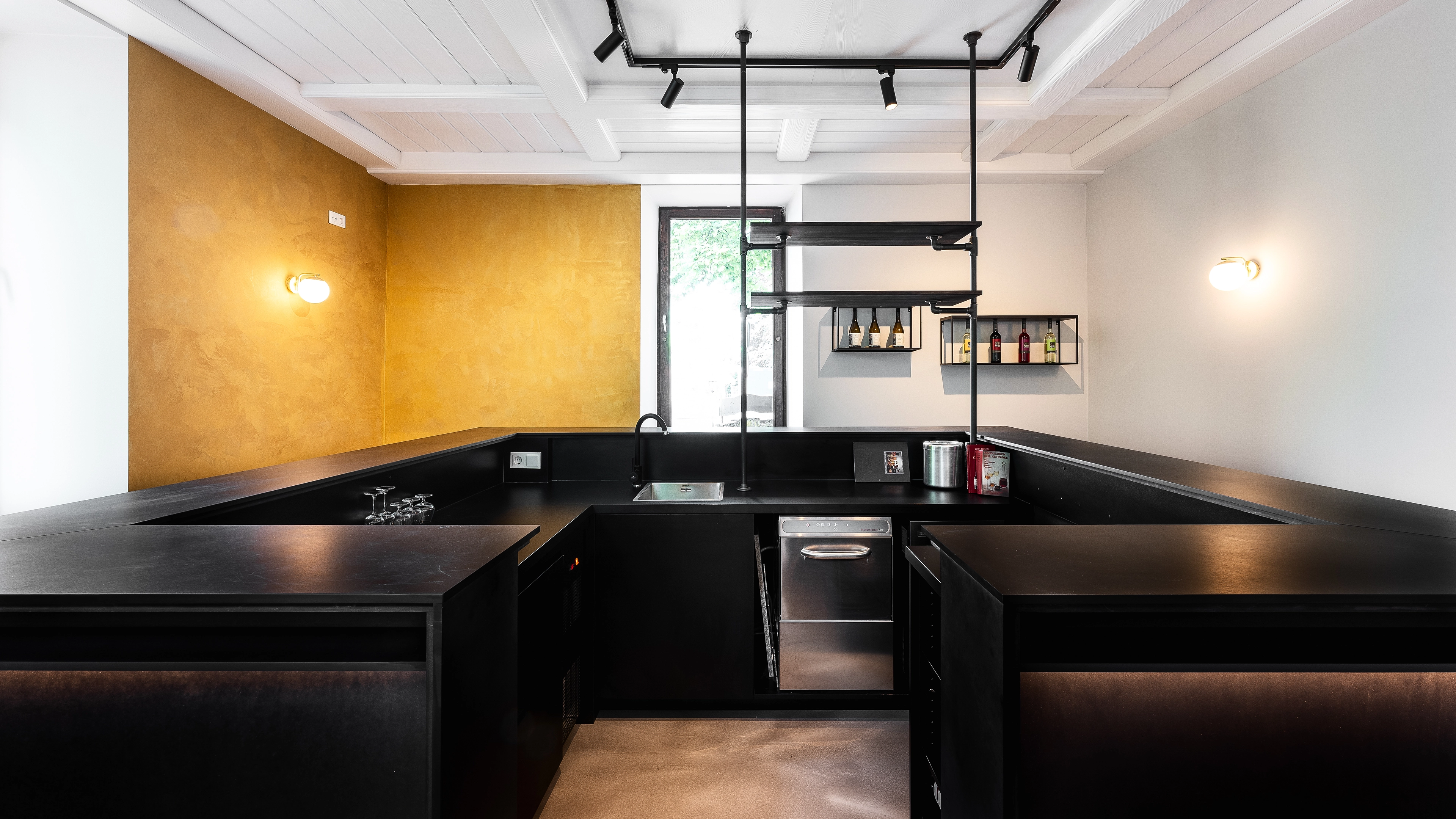 Modern black kitchen with an L-shaped countertop, a small sink, overhead shelves holding bottles, and yellow accent wall on the left.