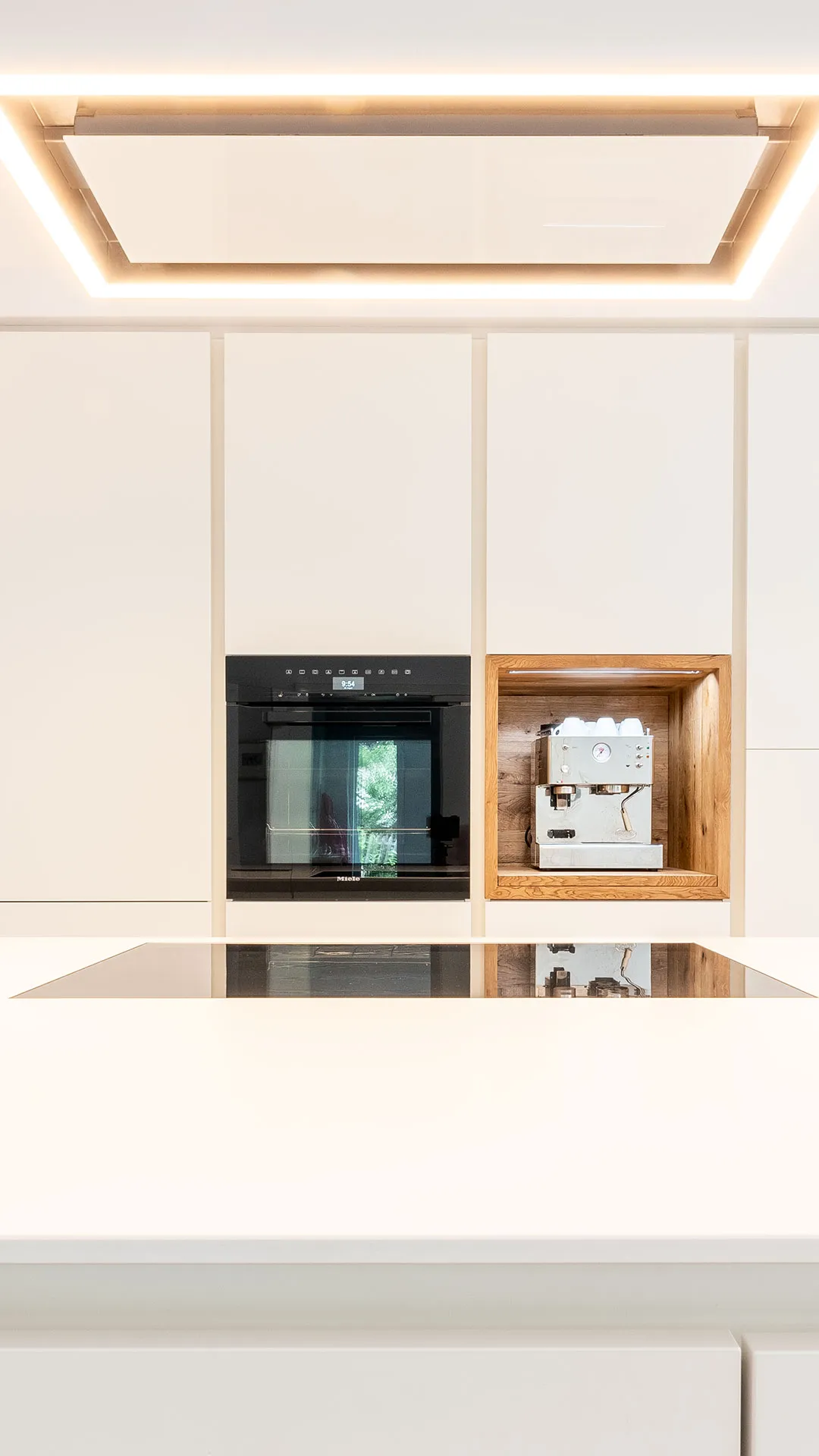 Modern kitchen wall with built-in black oven and wooden niche housing a white espresso machine.