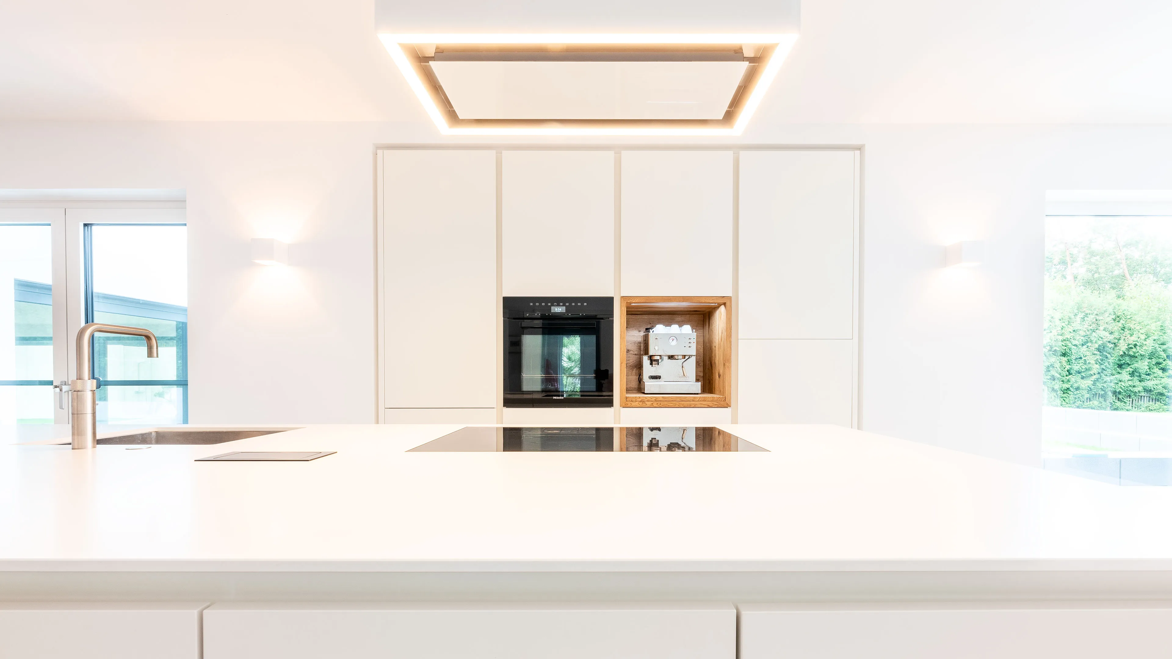 Minimalist white kitchen with a large island, built-in oven, and a coffee machine set in a wooden niche.