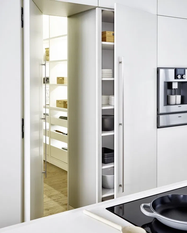 Modern white kitchen with a partially open pantry door revealing shelves with kitchenware and wooden boxes, and a built-in coffee machine on the wall.
