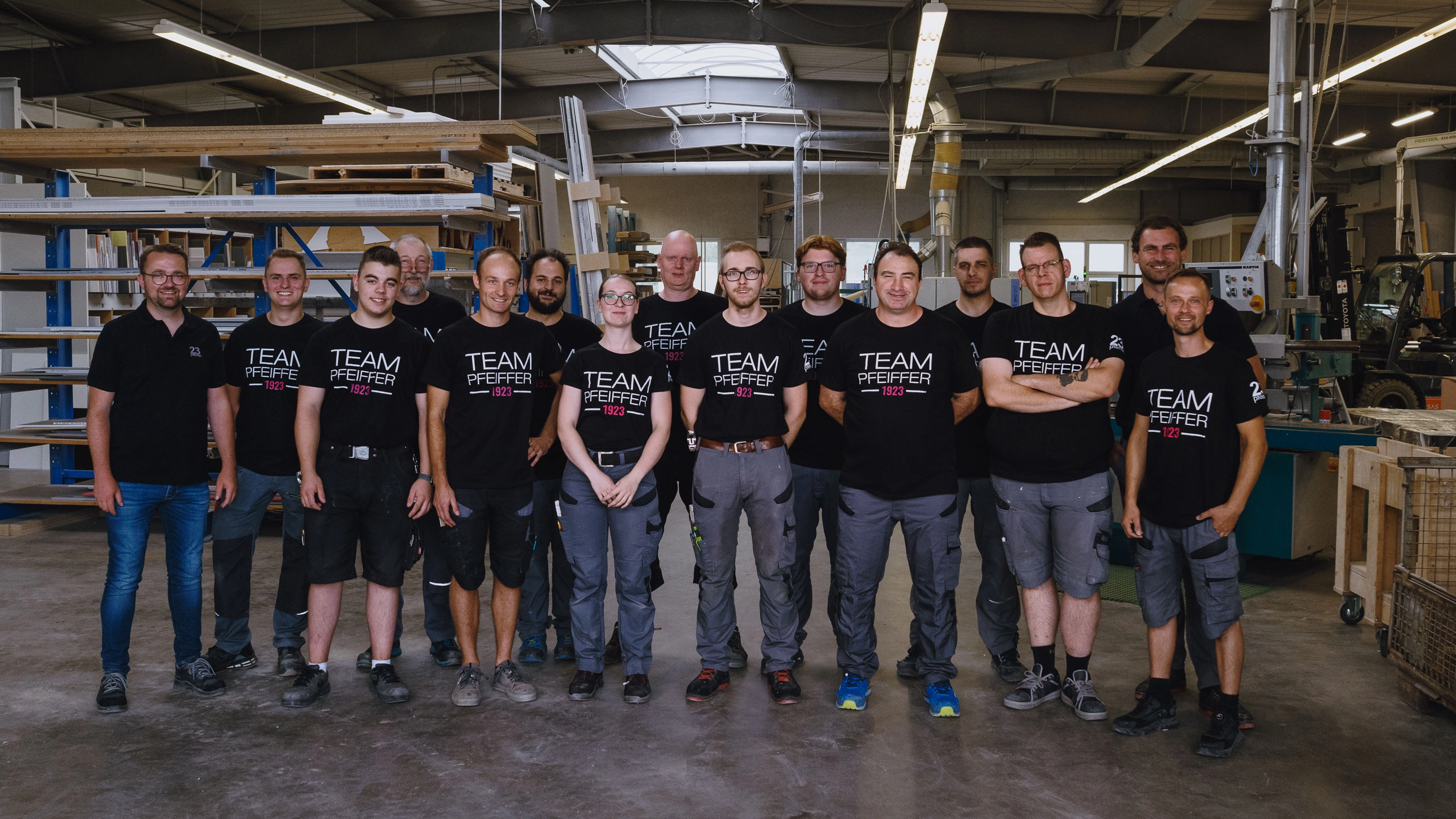 Group of fourteen people standing inside a workshop wearing black Team Pfeiffer 1923 shirts.