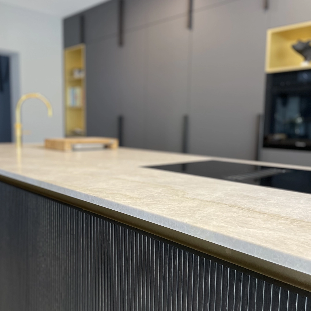 Close-up of a modern kitchen island with a beige marble countertop, a black cooktop, and dark grooved cabinetry, with blurred background cabinets and a gold faucet.