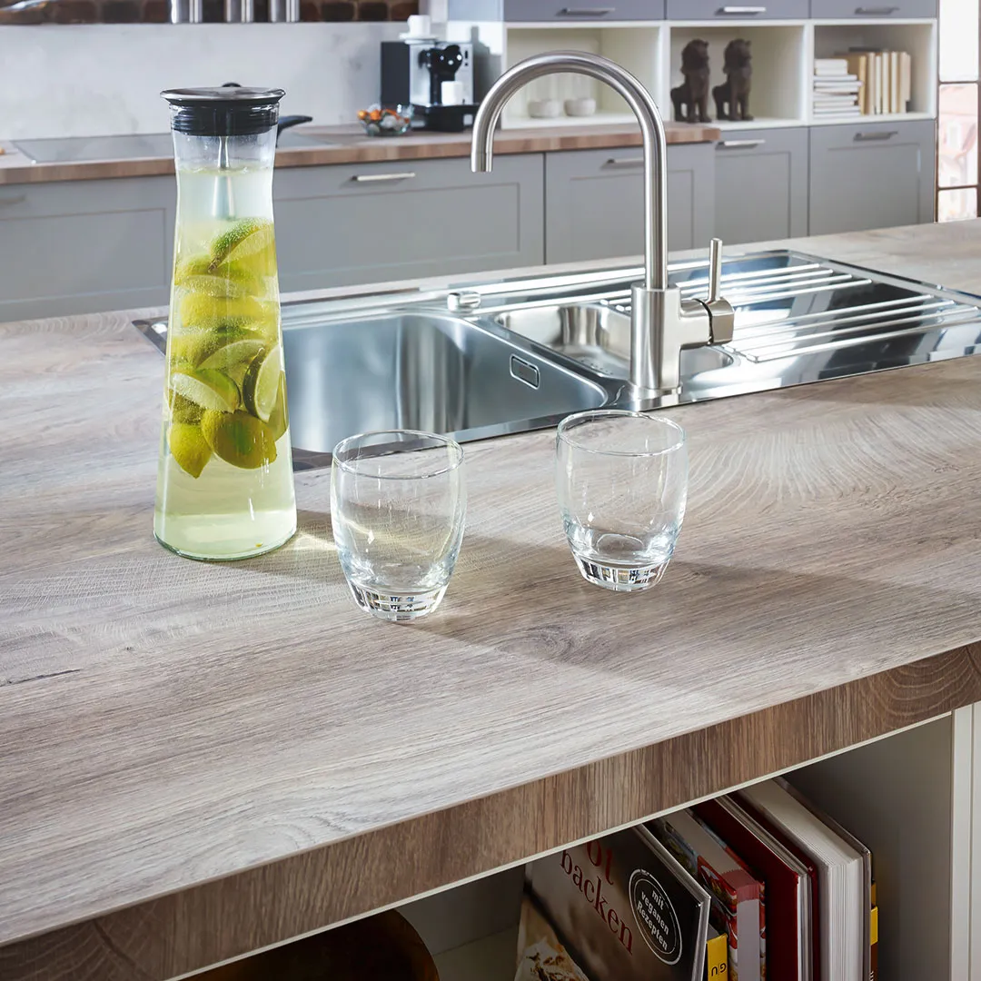 Modern kitchen sink with stainless steel faucet on wooden countertop, beside a glass pitcher with lime water and two empty glasses.