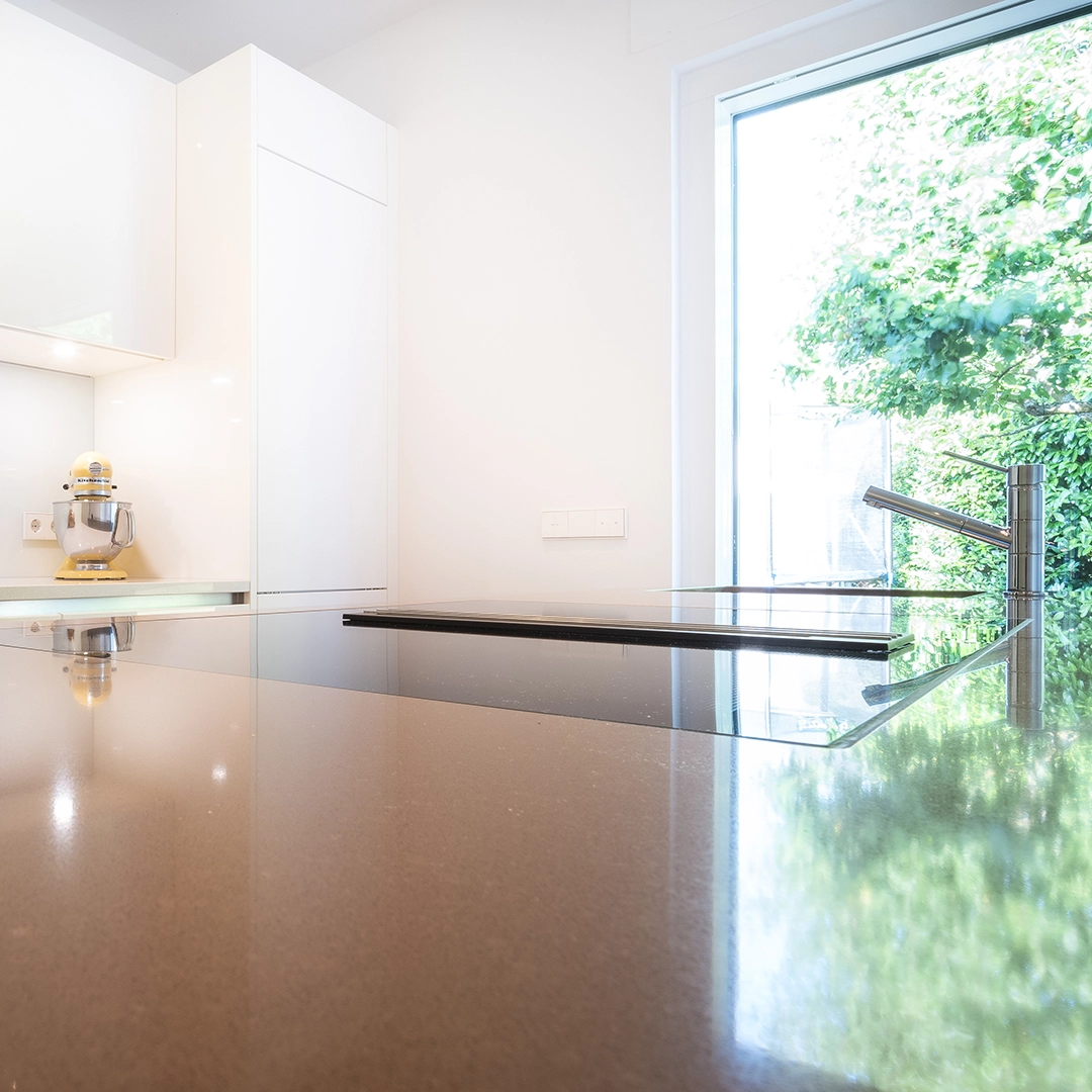 Modern kitchen countertop with built-in stovetop and stainless steel faucet next to a large window with green trees outside.