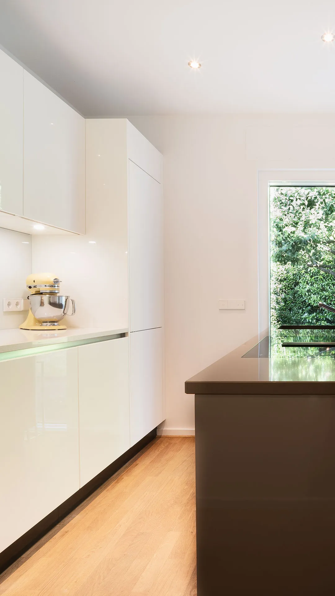 Modern kitchen with white cabinets, a yellow stand mixer on the counter, wooden flooring, and a window showing green foliage outside.