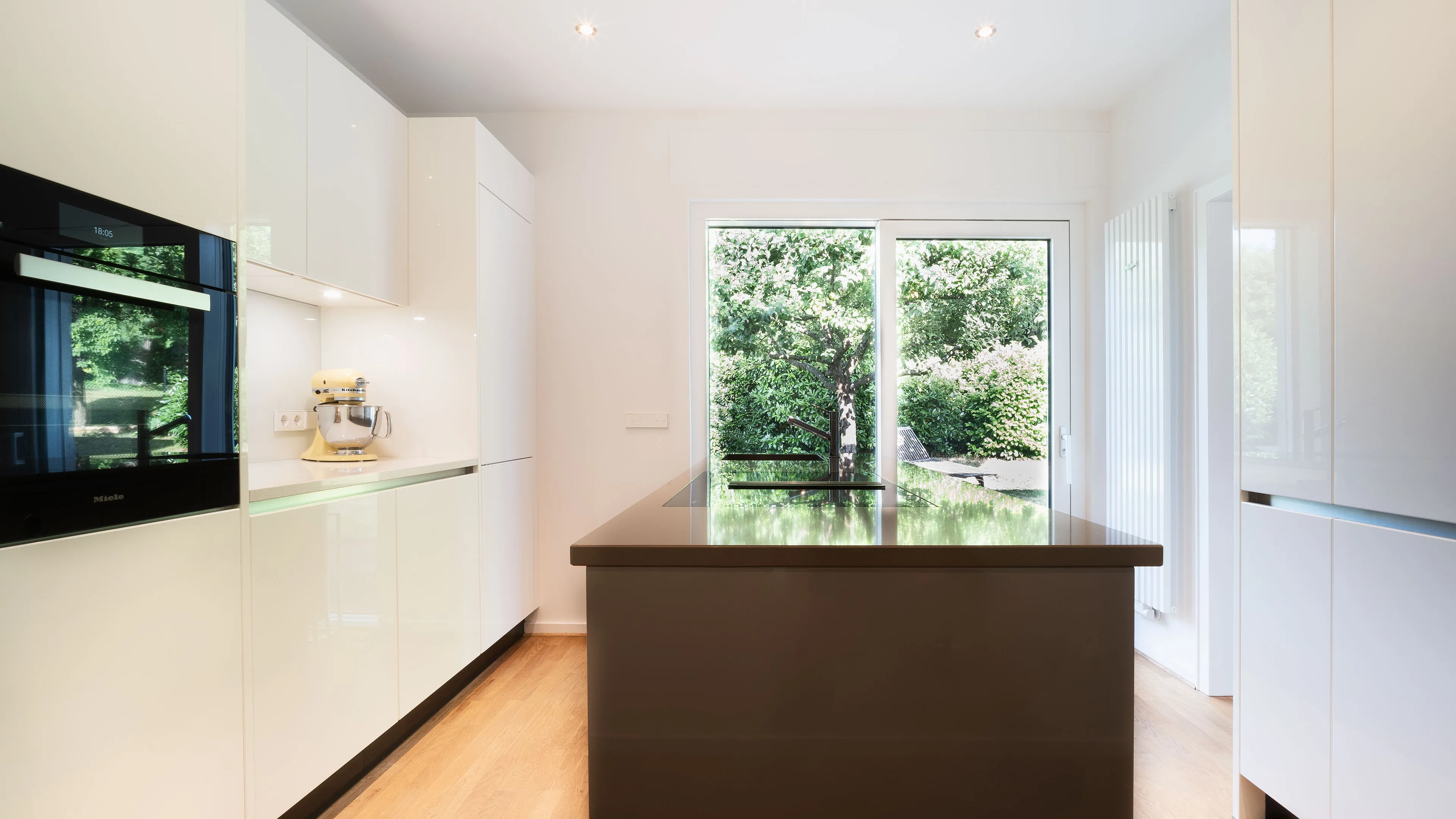 Modern kitchen with glossy white cabinets, a central island with cooktop and sink, wooden floor, and large window overlooking green garden.