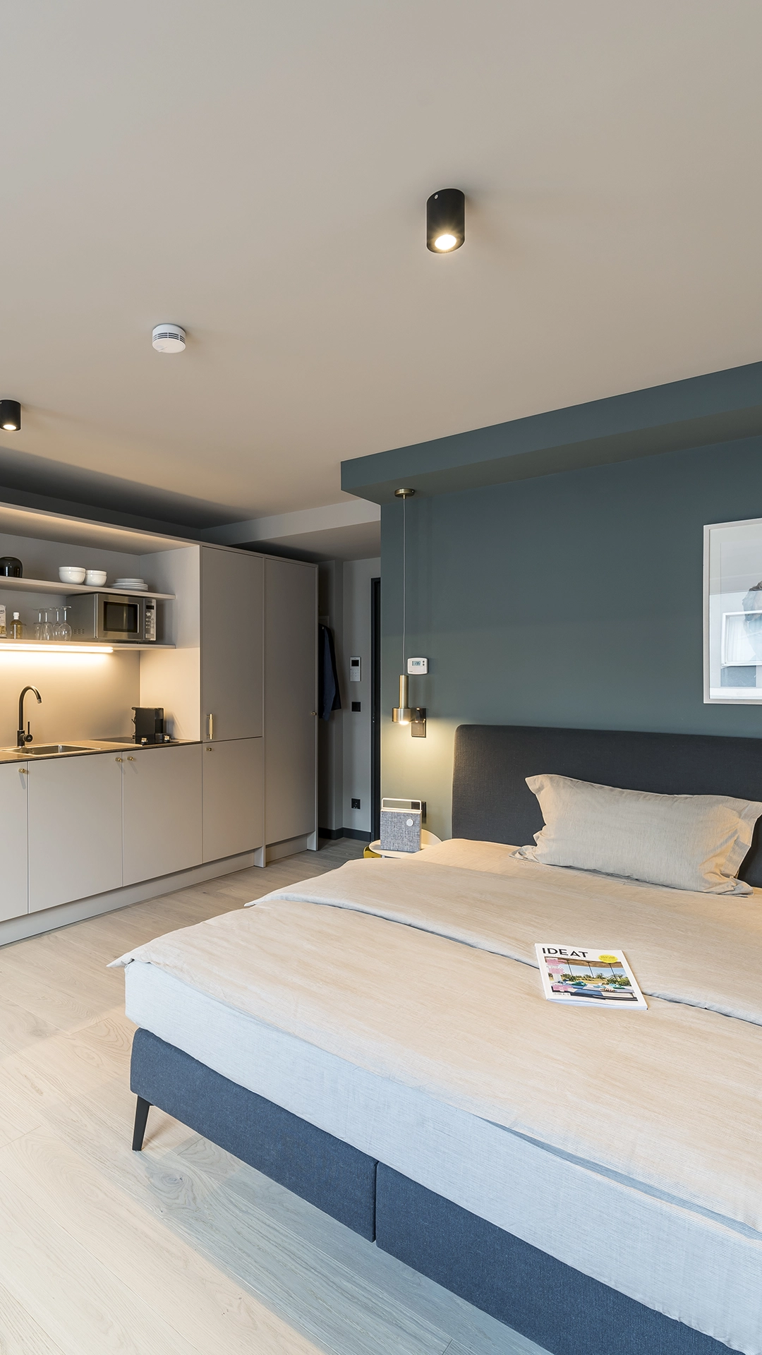 Modern studio apartment with a neatly made bed, beige bedding, blue upholstered headboard, and a small kitchen area with cabinets and a sink.