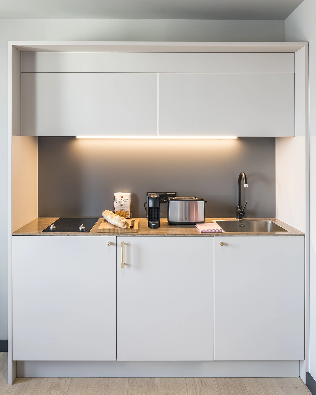Compact modern kitchen with white cabinets, electric stove, coffee machine, toaster, bread on a cutting board, and a sink with black faucet.