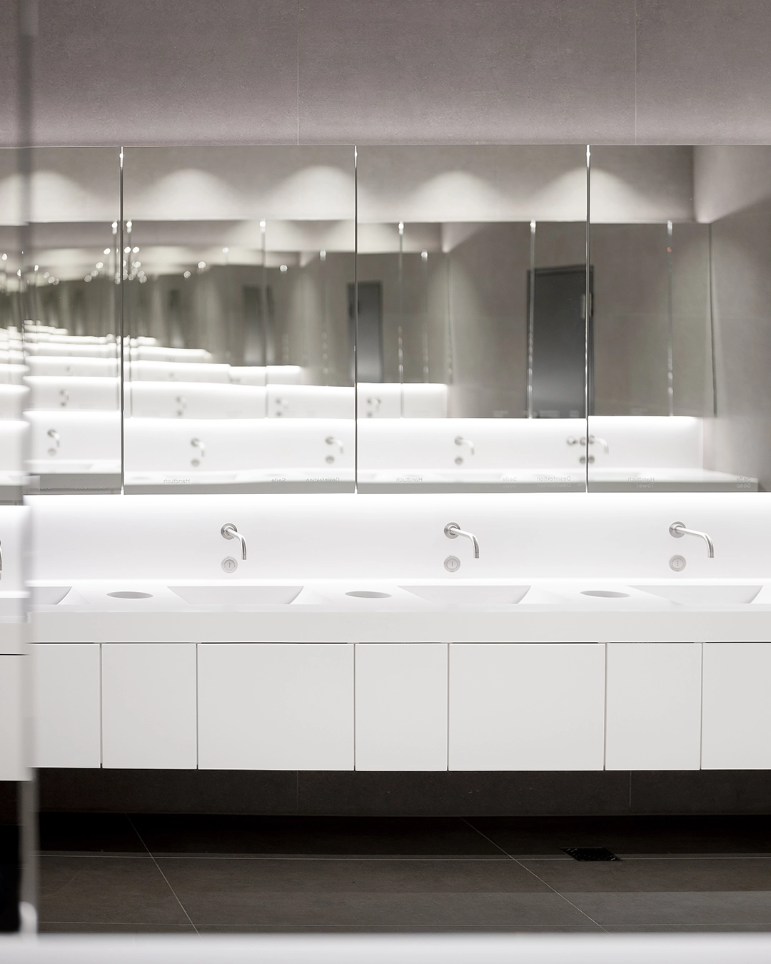 Modern public restroom with white sinks, stainless steel faucets, and a large mirror reflecting the room.
