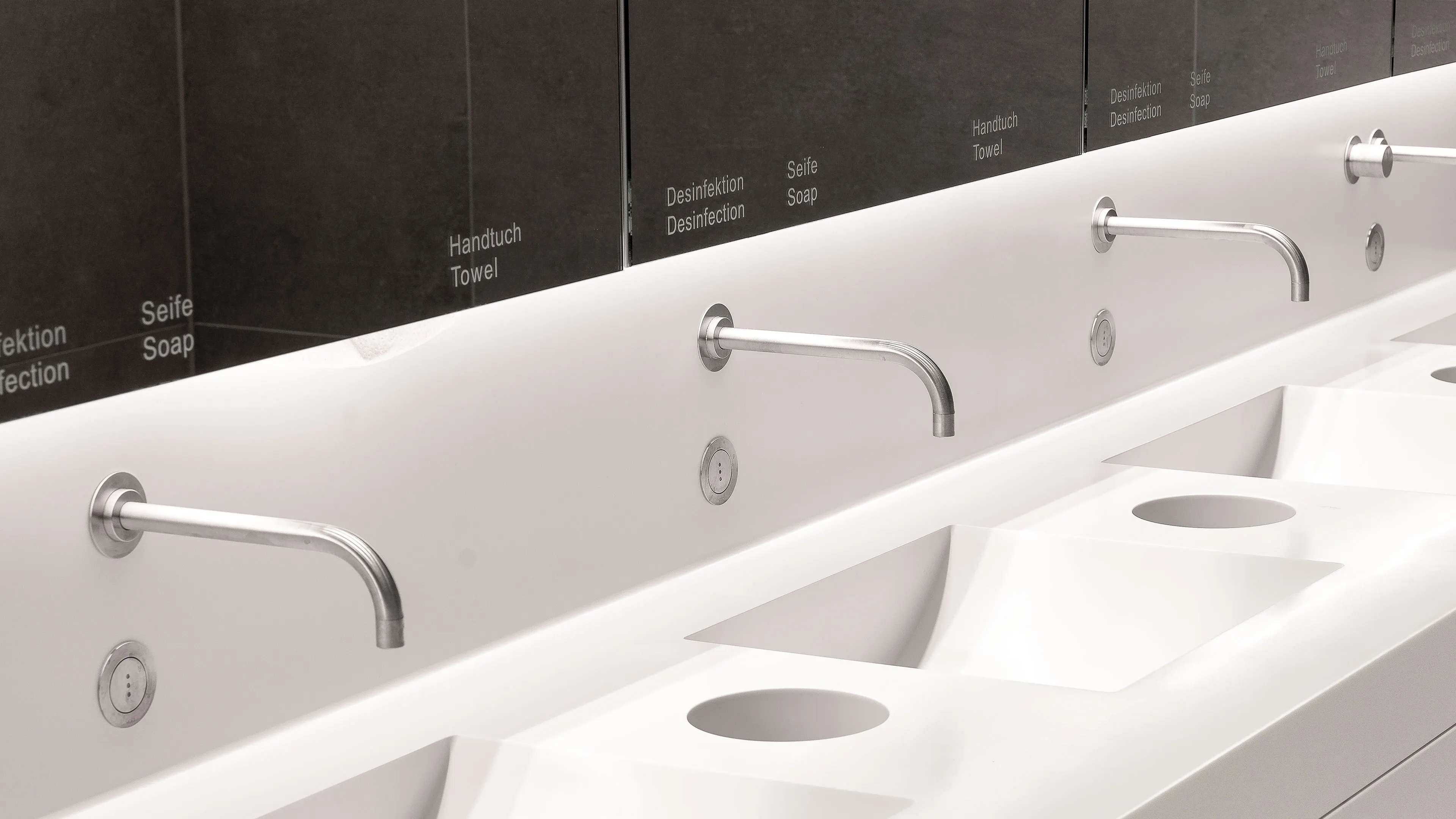 Modern white public restroom sinks with wall-mounted stainless steel faucets and mirrored backsplash labeled with soap, towel, and disinfection instructions.
