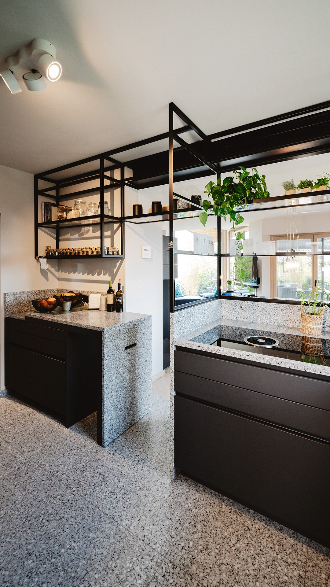 Modern kitchen with black cabinets, granite countertops and floor, open shelving with jars, and hanging green plants.