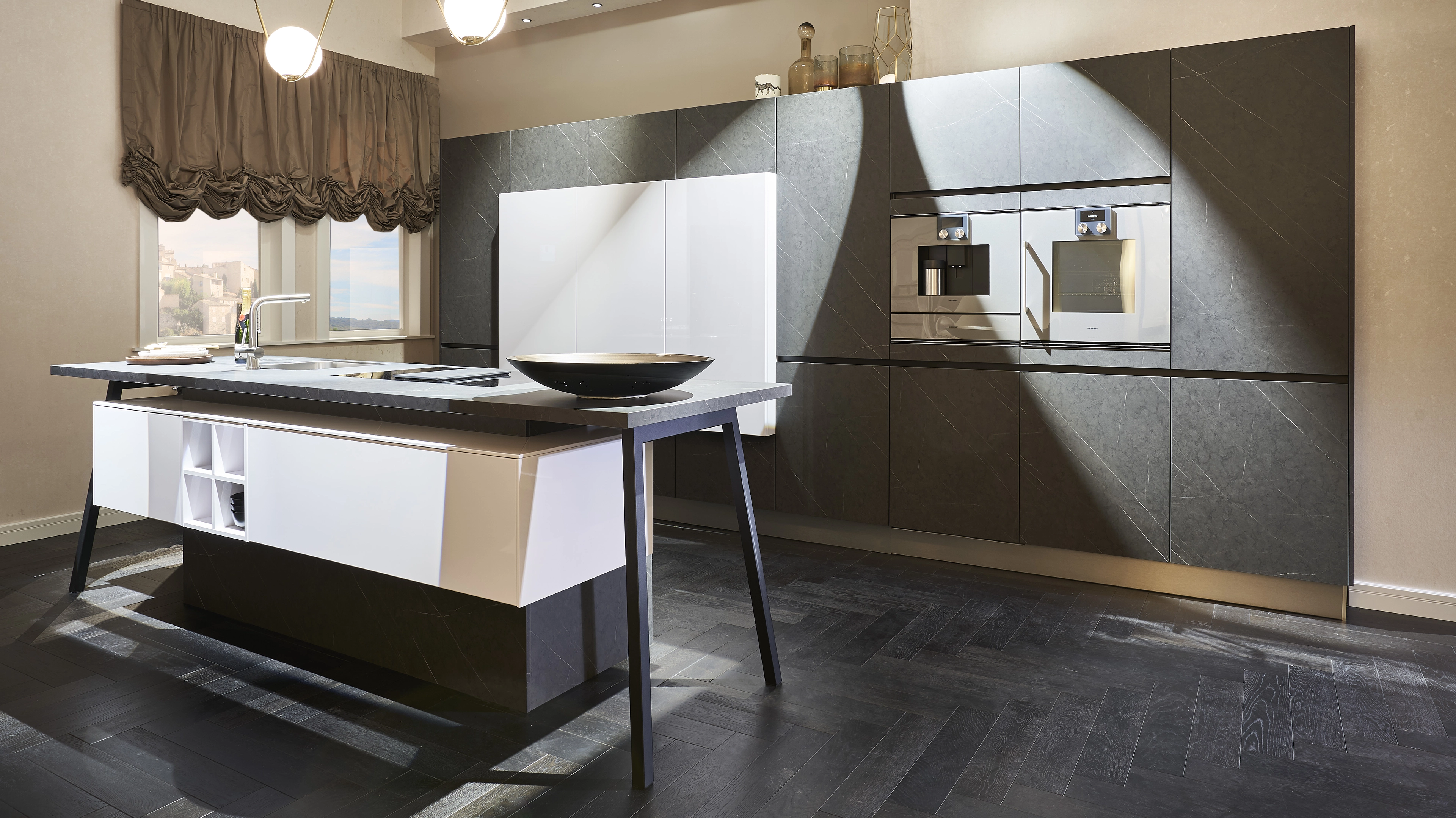 Modern kitchen with dark marble cabinets, built-in stainless steel oven and coffee machine, white island with black countertop and bowl, and window with brown curtains.