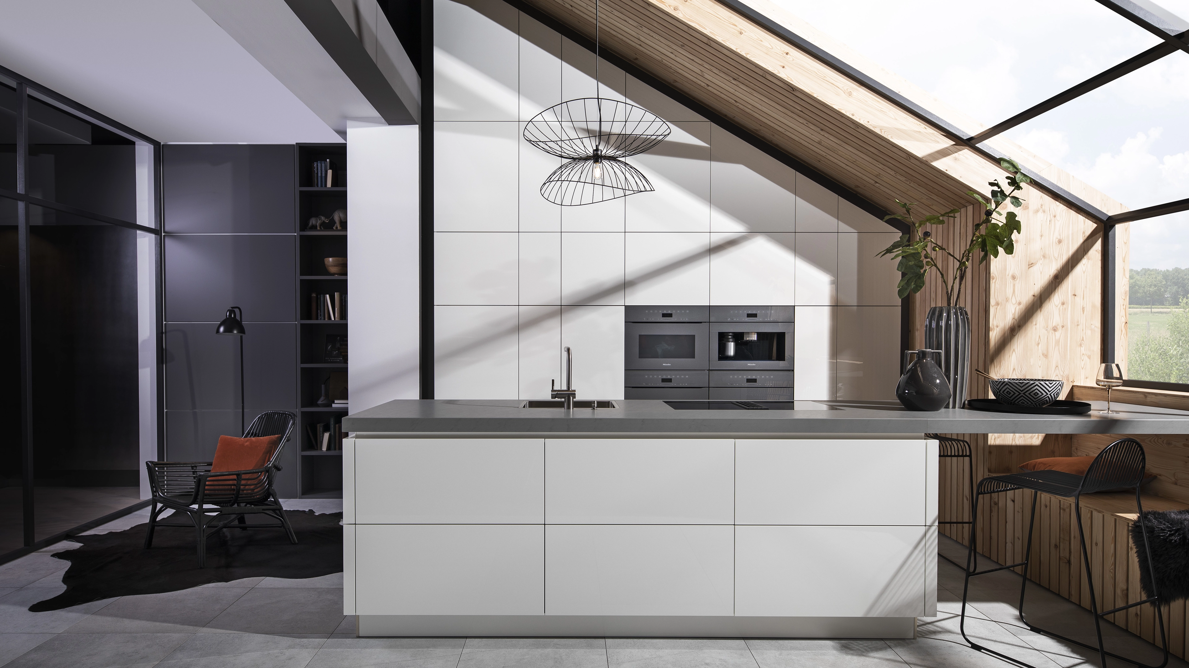 Modern kitchen with white cabinetry, gray countertop island, built-in ovens, black wireframe pendant light, and wooden slanted ceiling with large window.