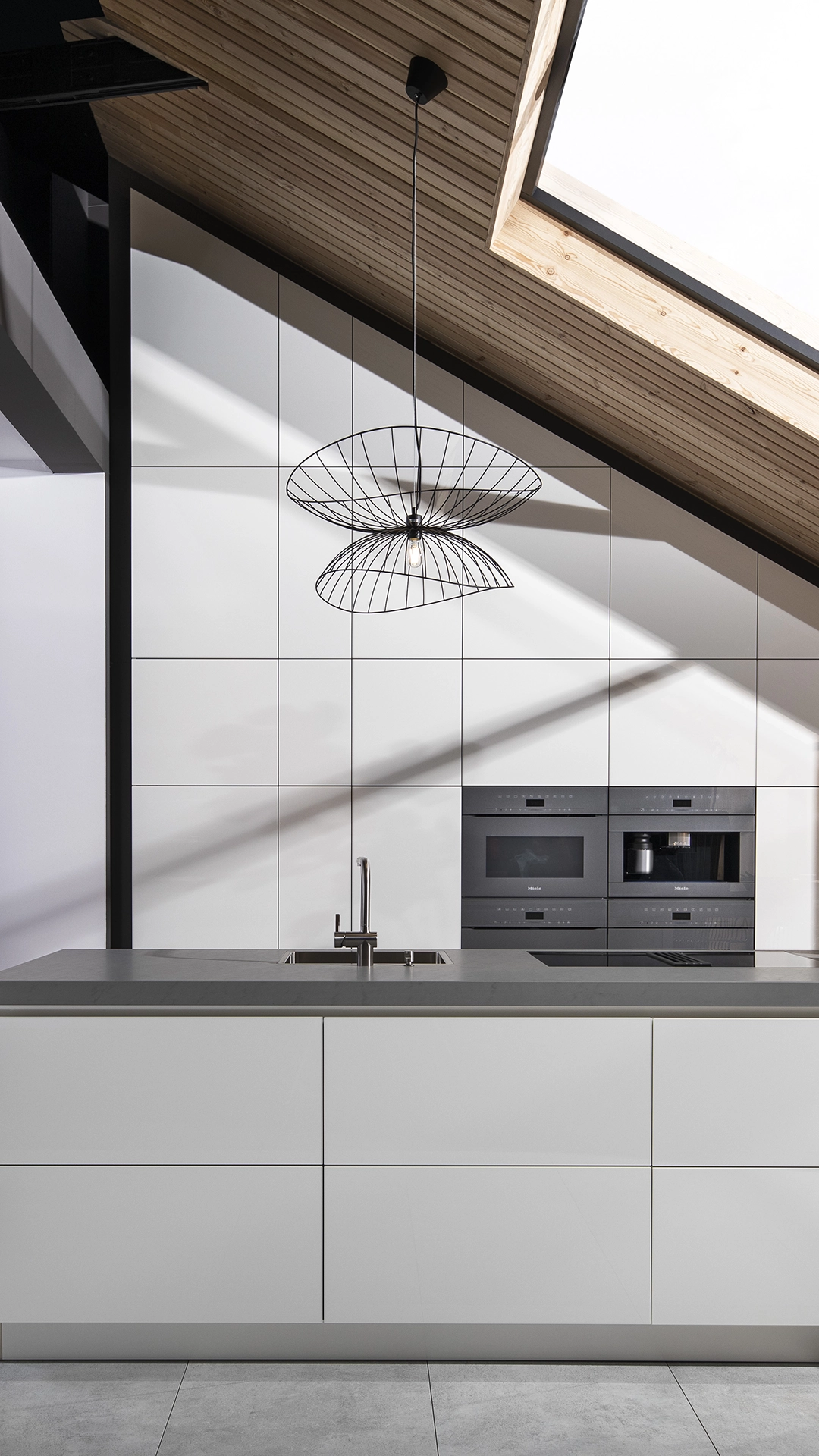 Modern kitchen with white cabinetry, gray countertop, built-in black appliances, wireframe pendant light, and angled wooden ceiling with skylight.