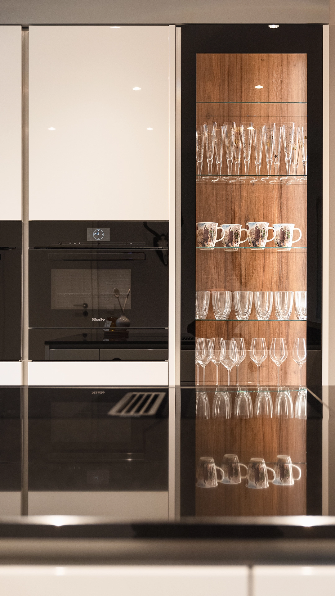 Modern kitchen cabinetry with built-in black oven and glass cabinet displaying glasses and mugs on wooden shelves.