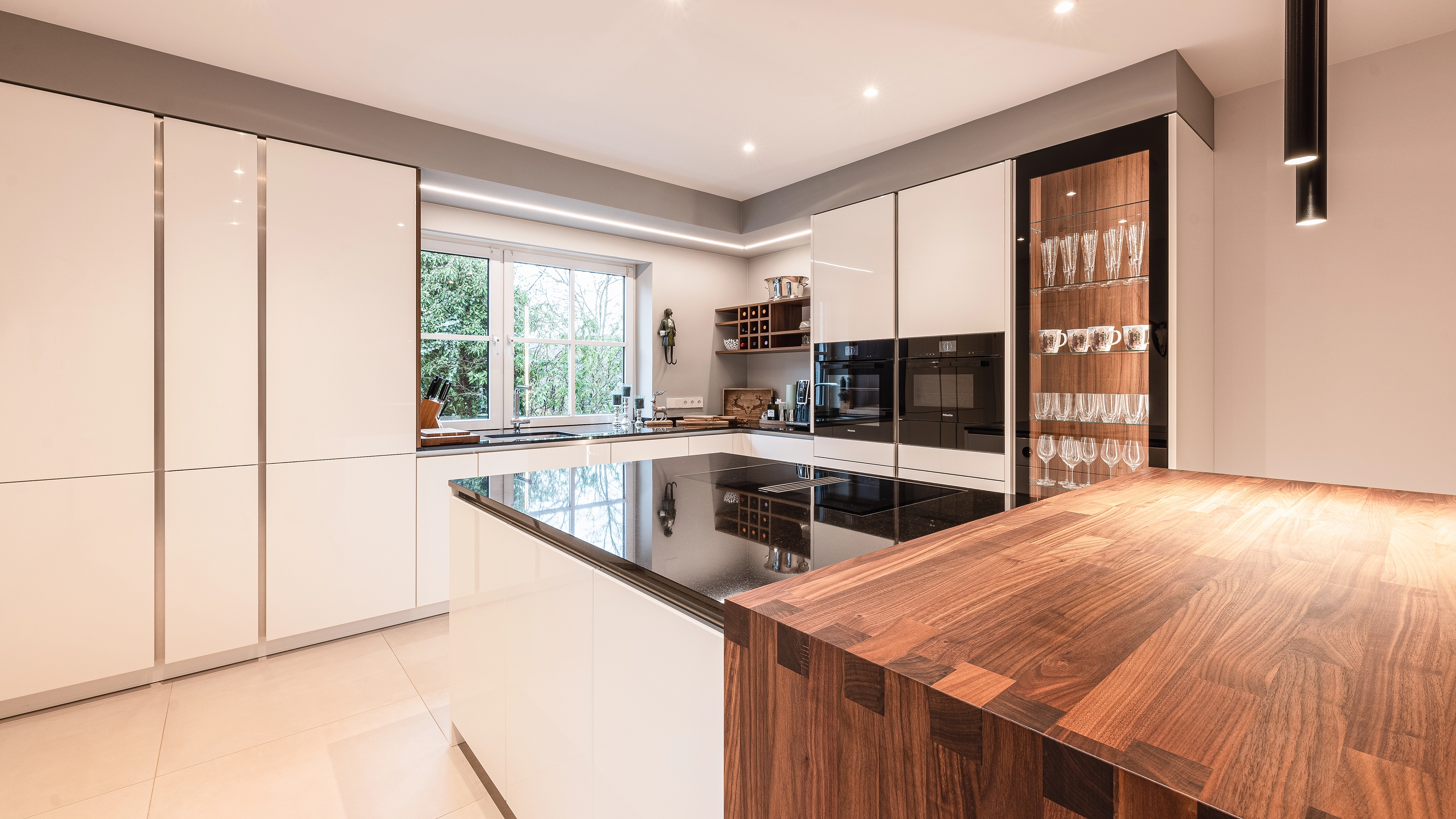 Modern kitchen with glossy white cabinets, black cooktop island, wooden countertop, built-in ovens, and a window overlooking greenery.