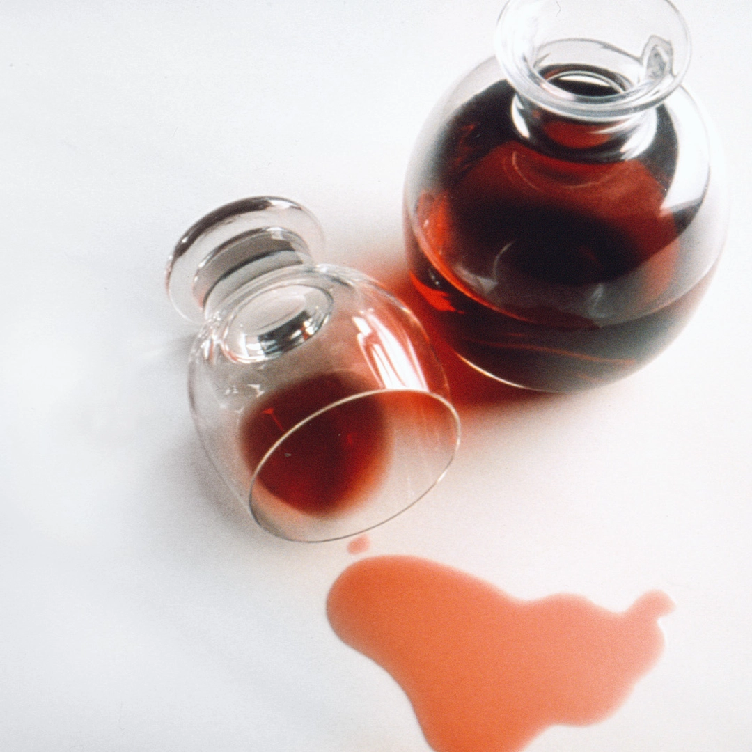 Glass decanter and overturned glass with red wine spilled on white surface.