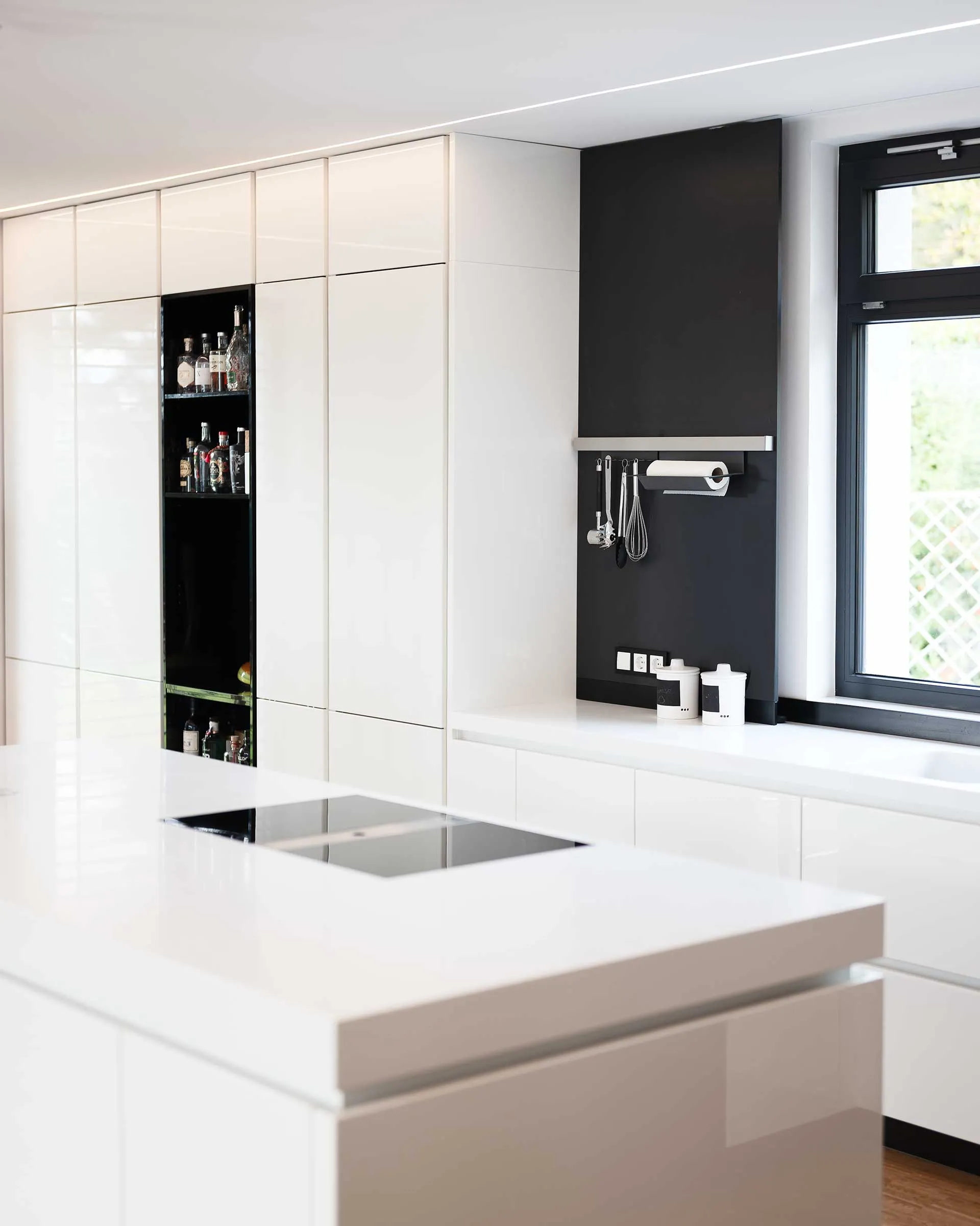 Modern kitchen with white glossy cabinets, a built-in stovetop on a white island, black open shelves with bottles, and a window next to a black wall with hanging utensils and paper towel holder.