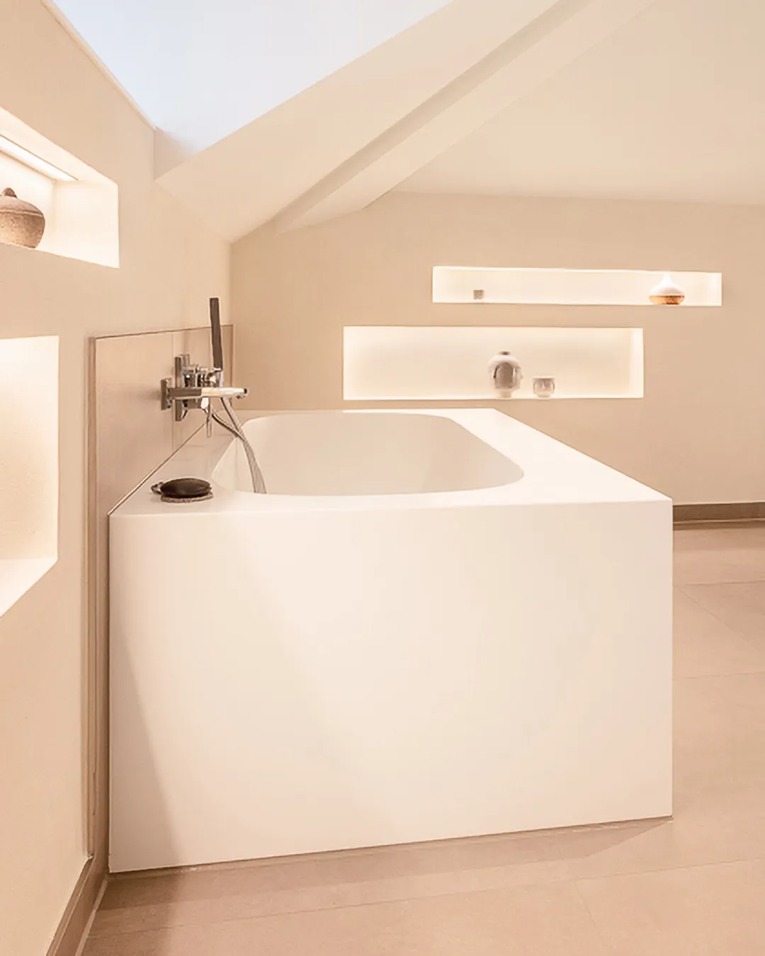Minimalist white bathtub with a modern faucet under a slanted ceiling in a softly lit bathroom with recessed shelves.