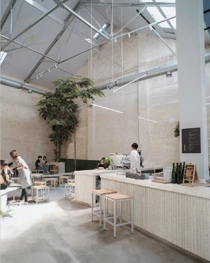 Bright modern café interior with high ceilings, white brick walls, minimalist furniture, and a barista behind the counter.