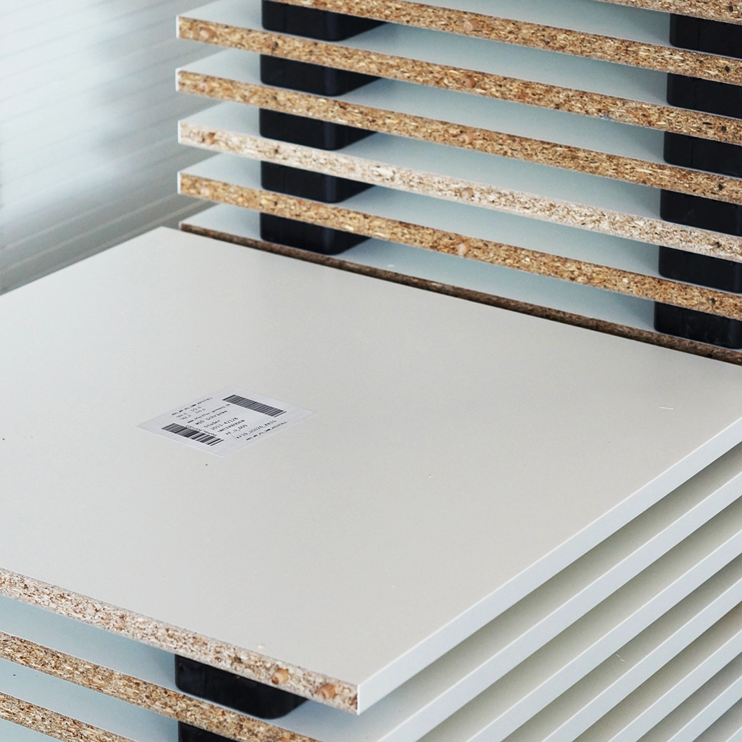Stack of laminated particleboard panels with white surfaces and exposed chipboard edges, one panel featuring a label sticker.