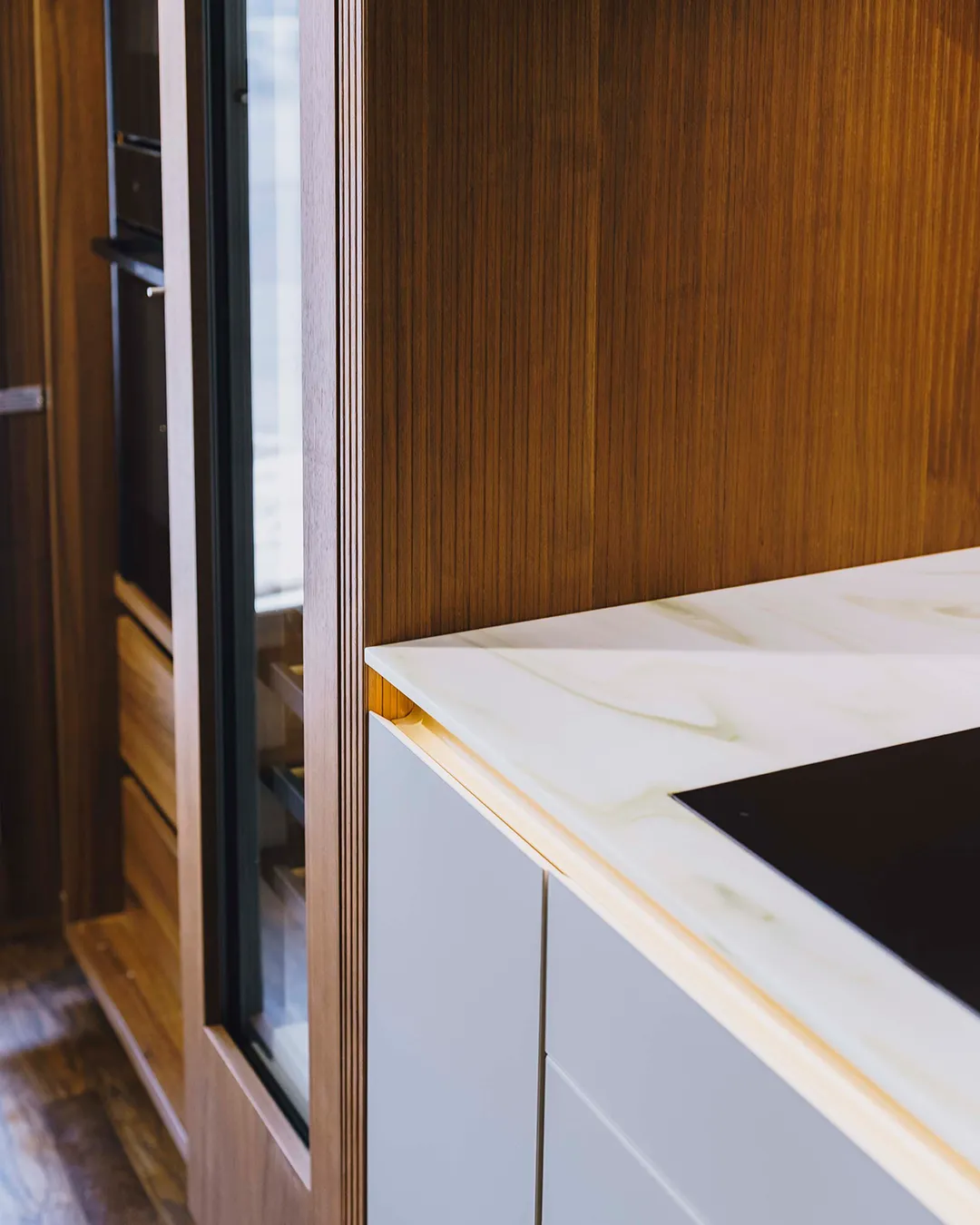 Modern kitchen corner featuring white marble countertop, black cooktop, and warm wood cabinetry with vertical grain.