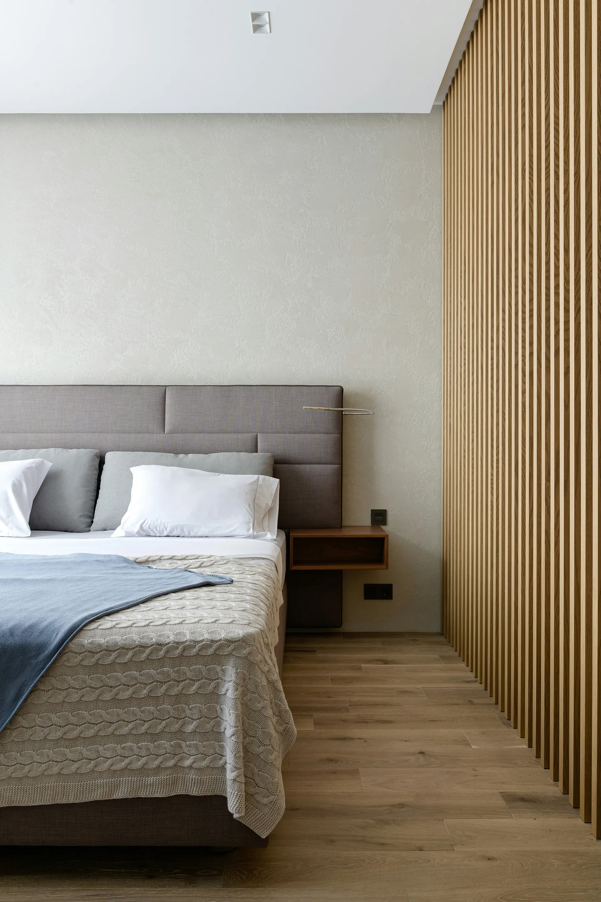 Modern bedroom with a gray upholstered bed, textured beige blanket, blue throw, and wooden slatted wall paneling.