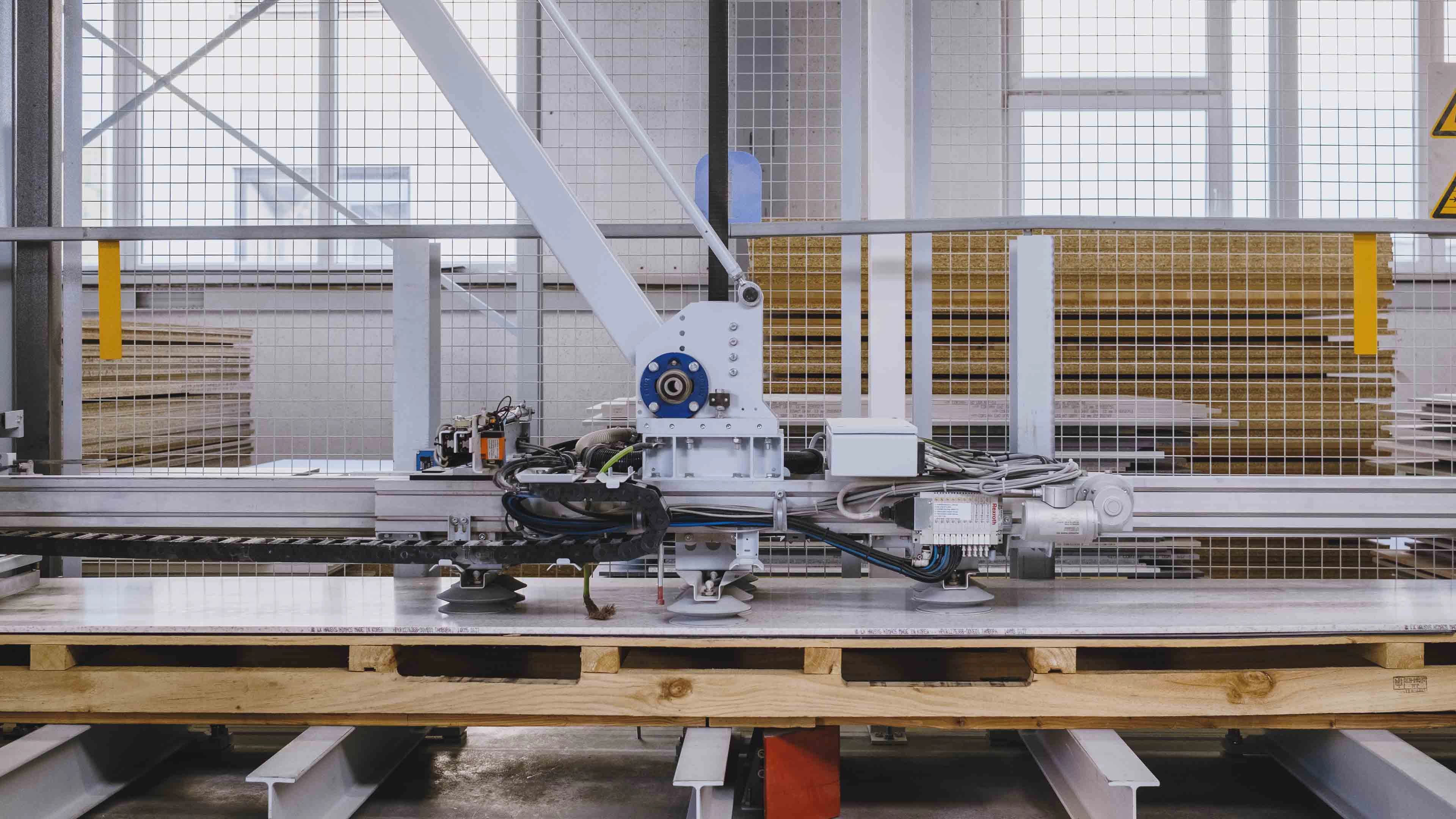 Industrial machine with suction cups lifting a sheet of material on a wooden pallet inside a factory.