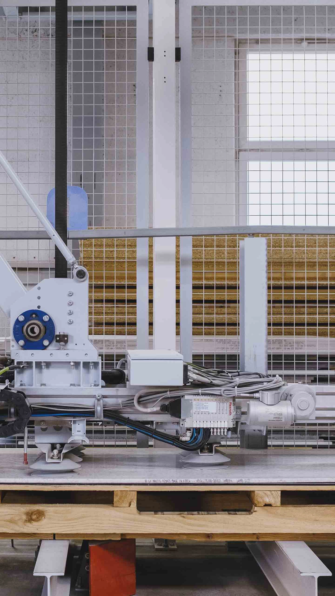 Industrial robotic arm mounted on a wooden platform inside a factory with stacked wooden boards in the background.
