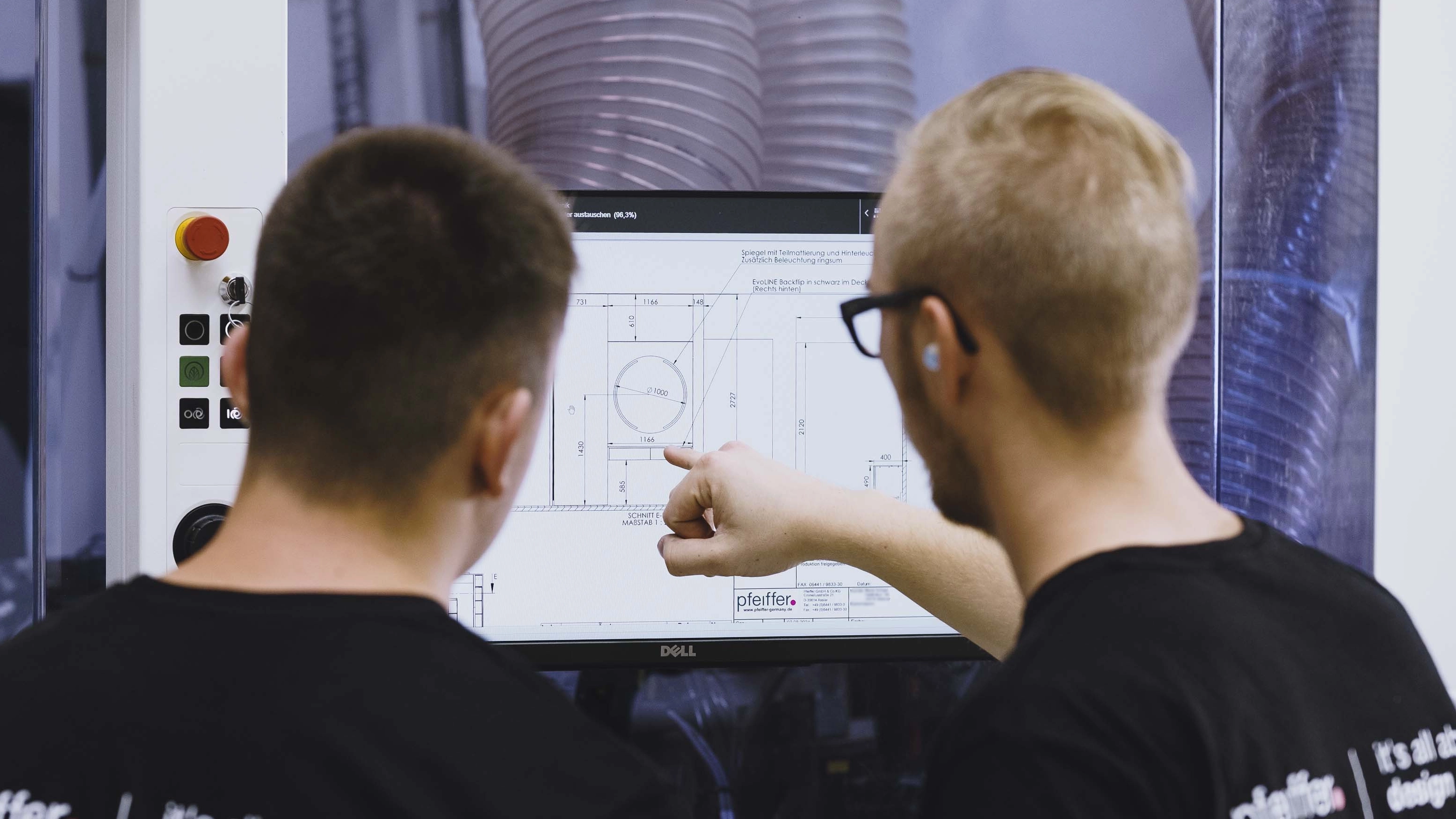 Two men in black shirts examining a technical drawing on a Dell monitor, one pointing at the screen.