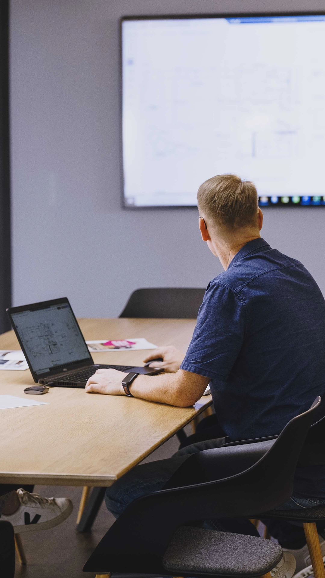 Man sitting at a table using a laptop and looking at a large screen with architectural or technical drawings.