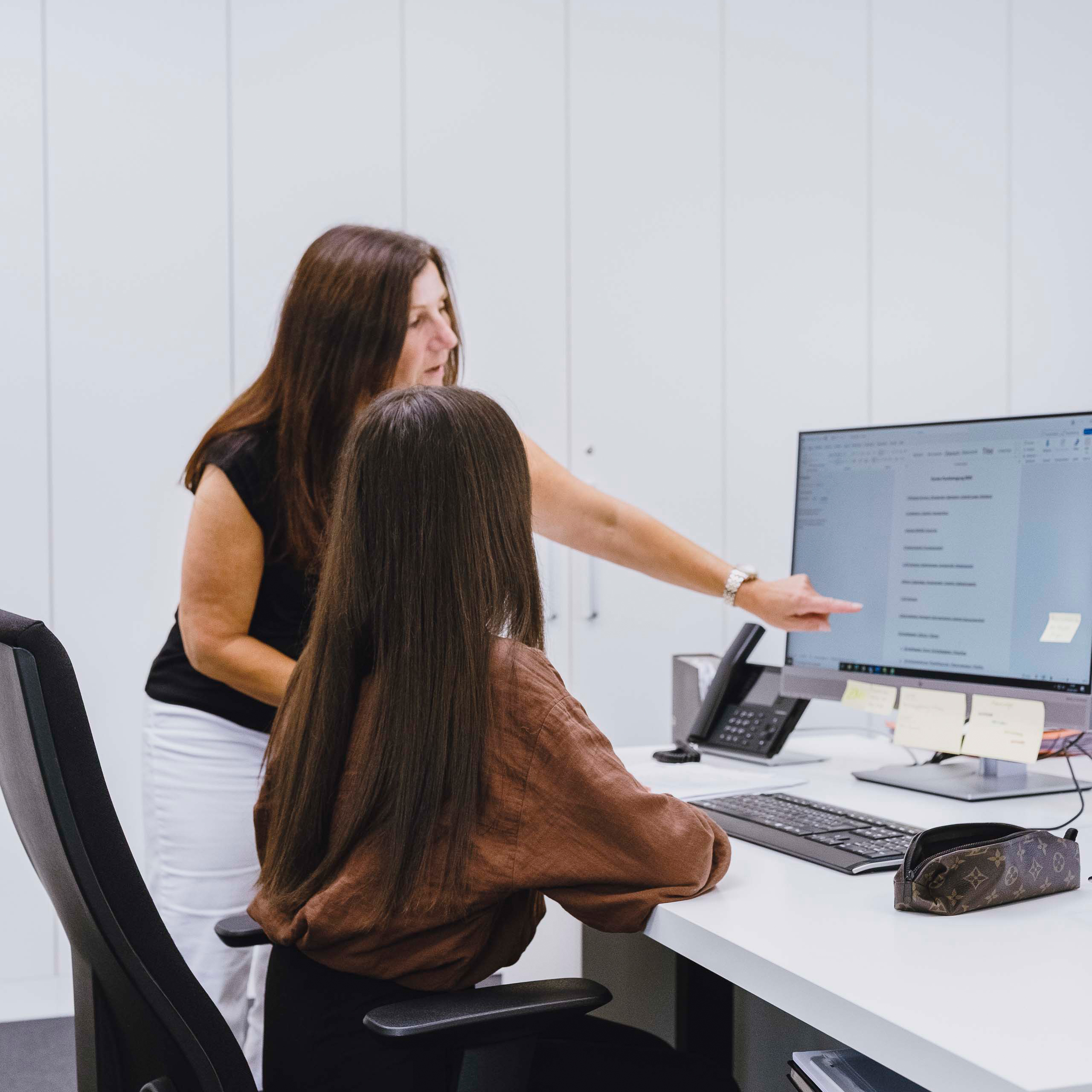 A woman standing and pointing at a computer screen while another woman is seated and looking at the monitor in a modern office.