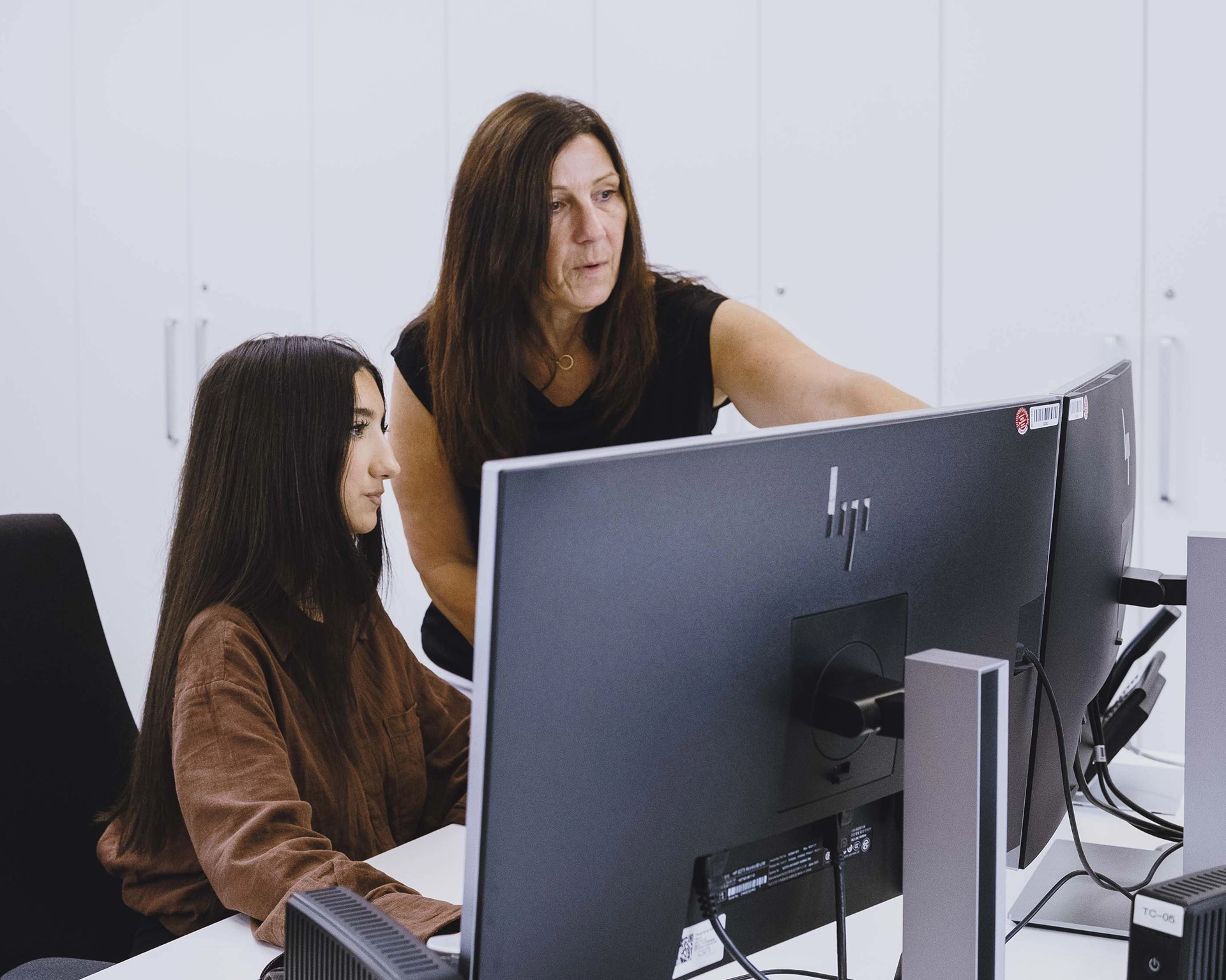 A woman standing and pointing at a computer screen while another woman is seated and looking at the monitor in a modern office.