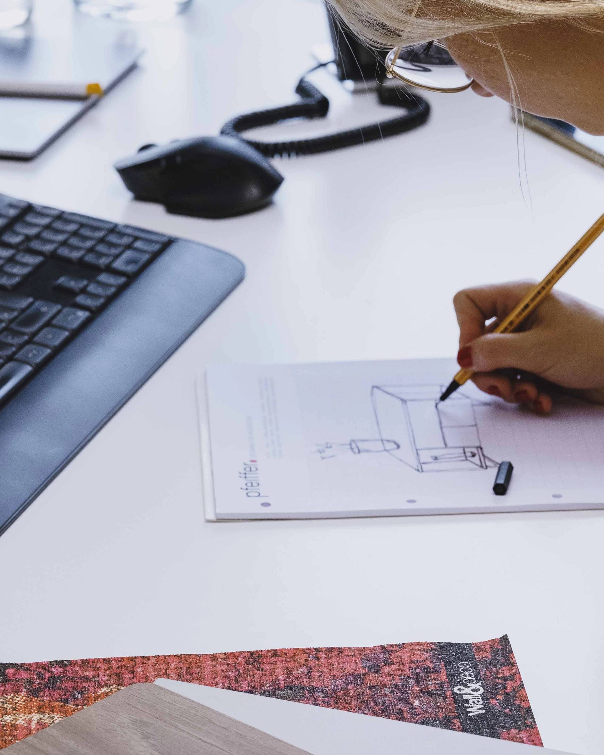 Person sketching a house design on paper at a desk with a keyboard, mouse, and material samples.