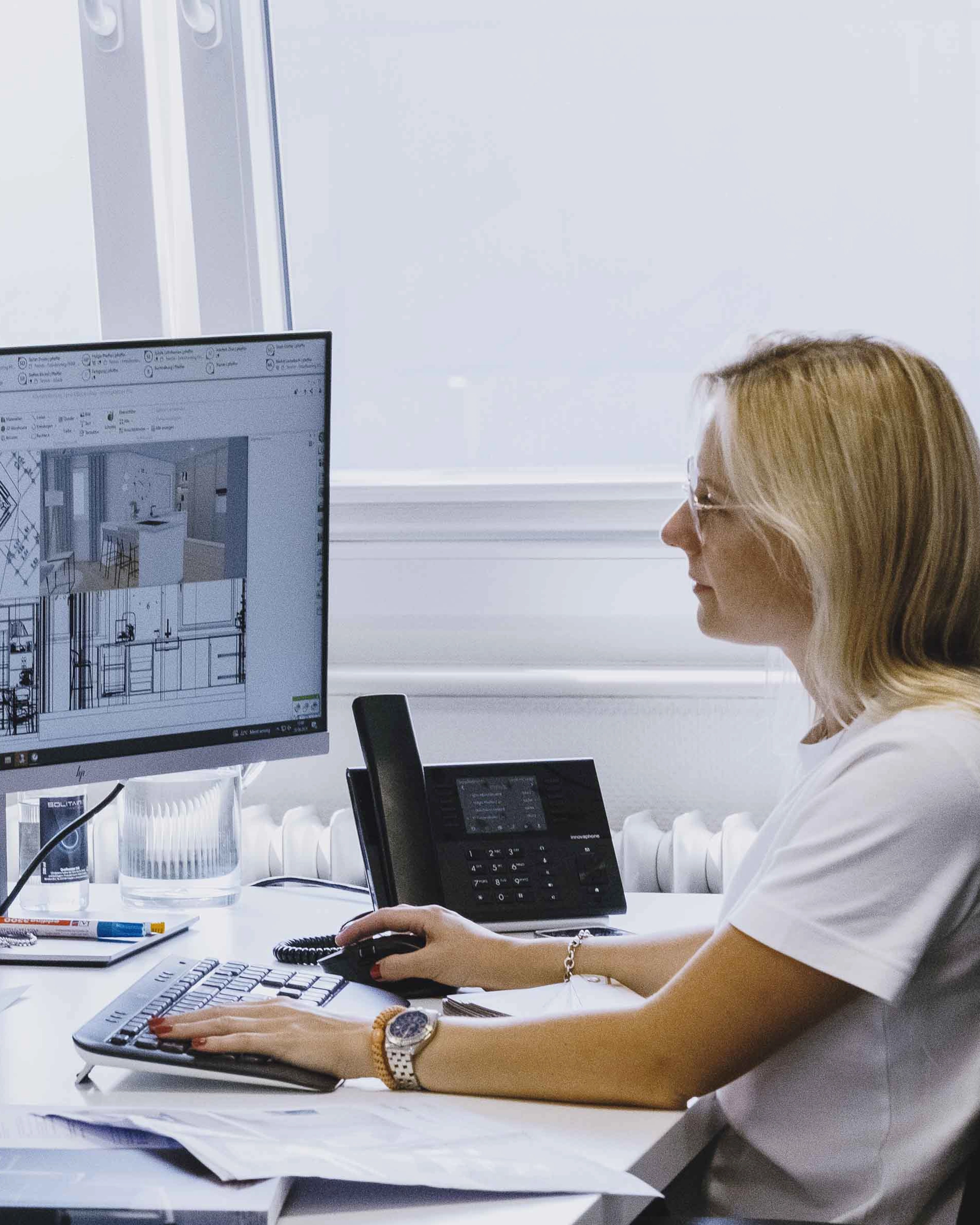 Woman with blonde hair and glasses working at a desk using a computer mouse and keyboard, viewing architectural designs on the monitor.