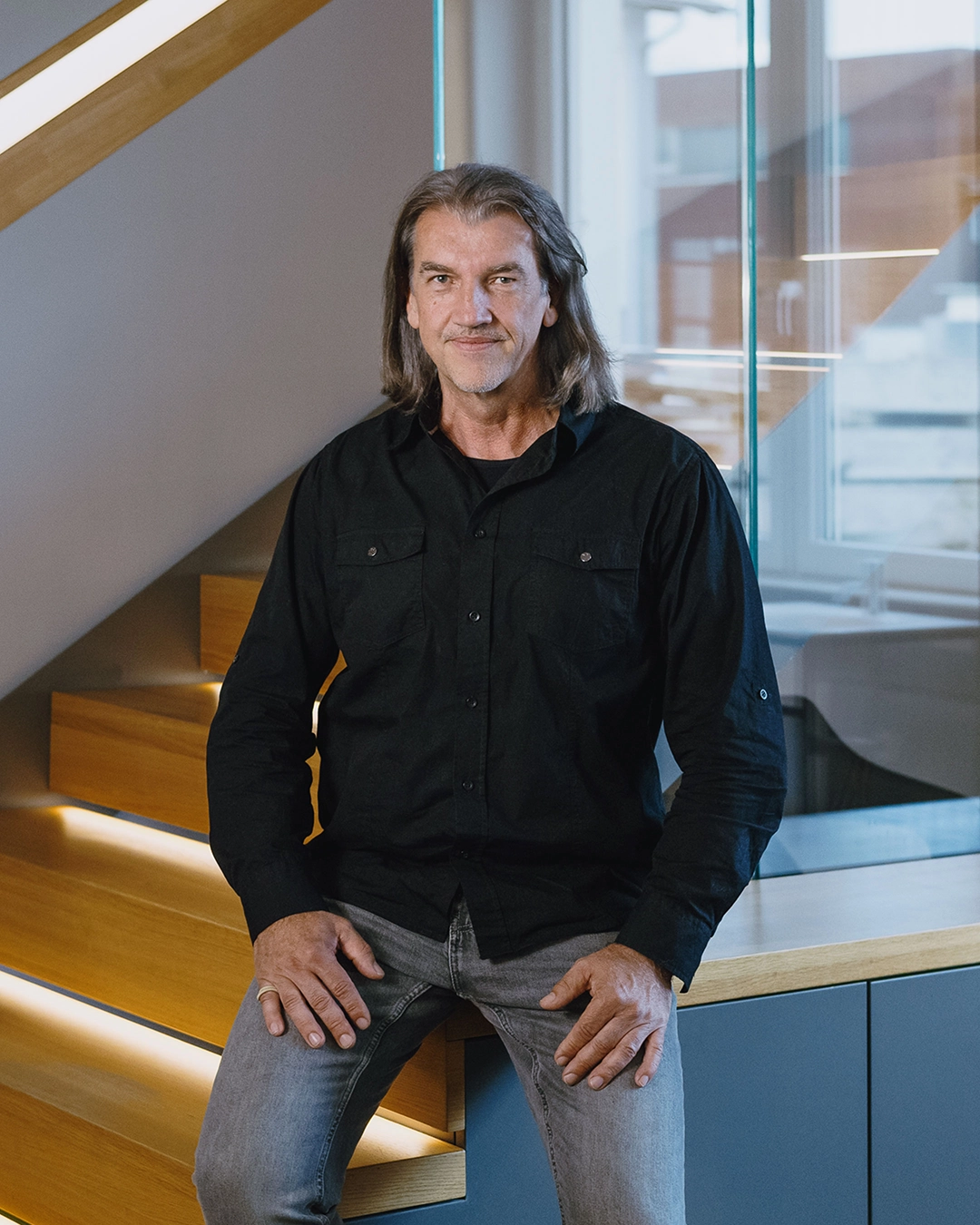 Middle-aged man with shoulder-length hair wearing a black shirt and gray jeans sitting on wooden stairs in a modern indoor setting.