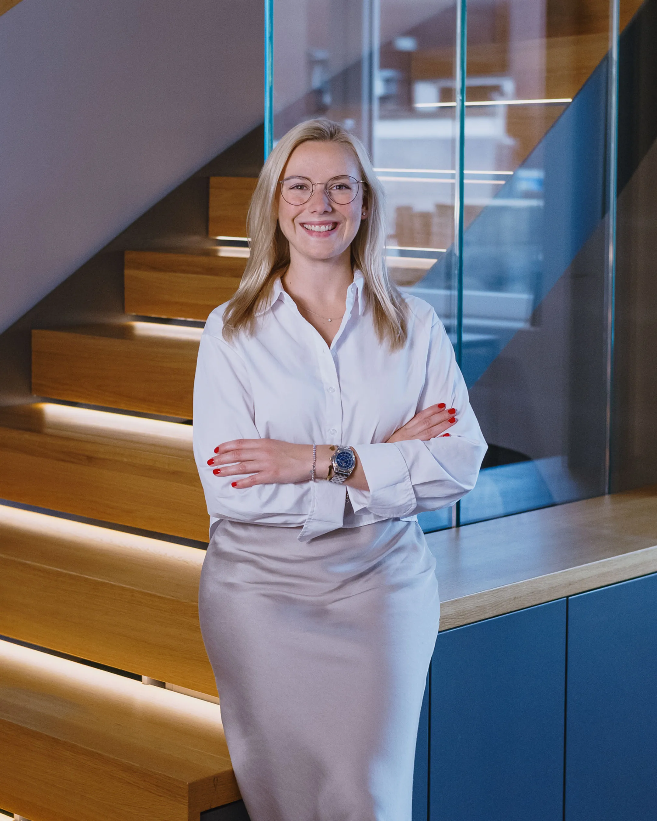 Smiling blonde woman with glasses, wearing a white blouse and gray skirt, standing with arms crossed in front of wooden stairs with glass panels.