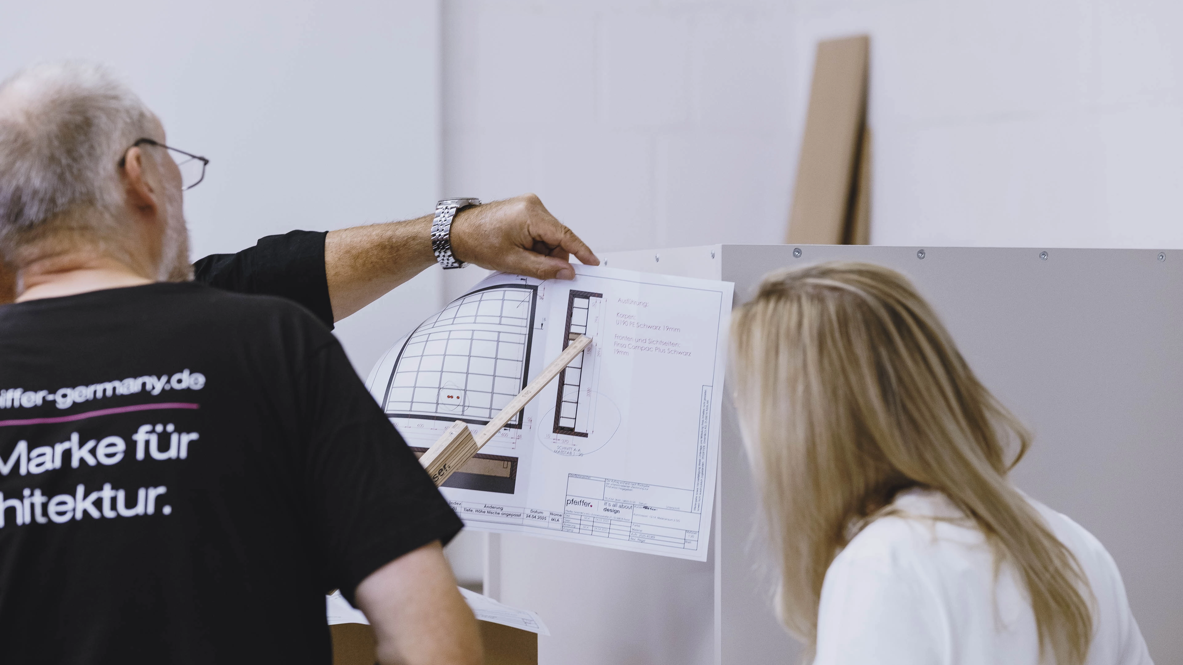 Man pointing at architectural blueprint with a wooden ruler while woman observes in a light-colored room.
