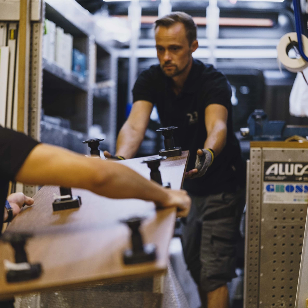 Two men carrying a large wooden board with black adjustable feet in a workshop.