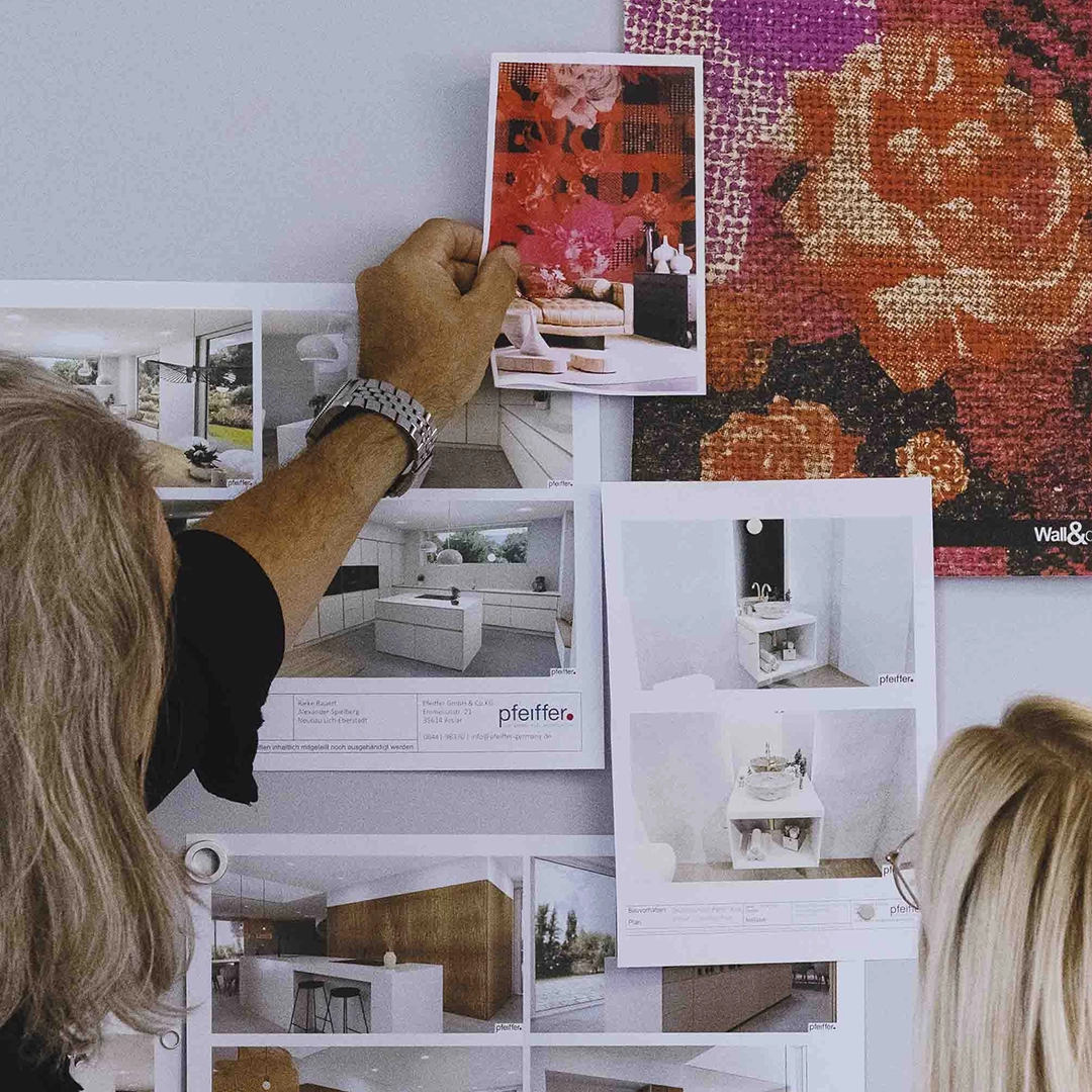 Two people reviewing interior design photos pinned on a wall, including modern kitchens and a vibrant floral wallpaper.