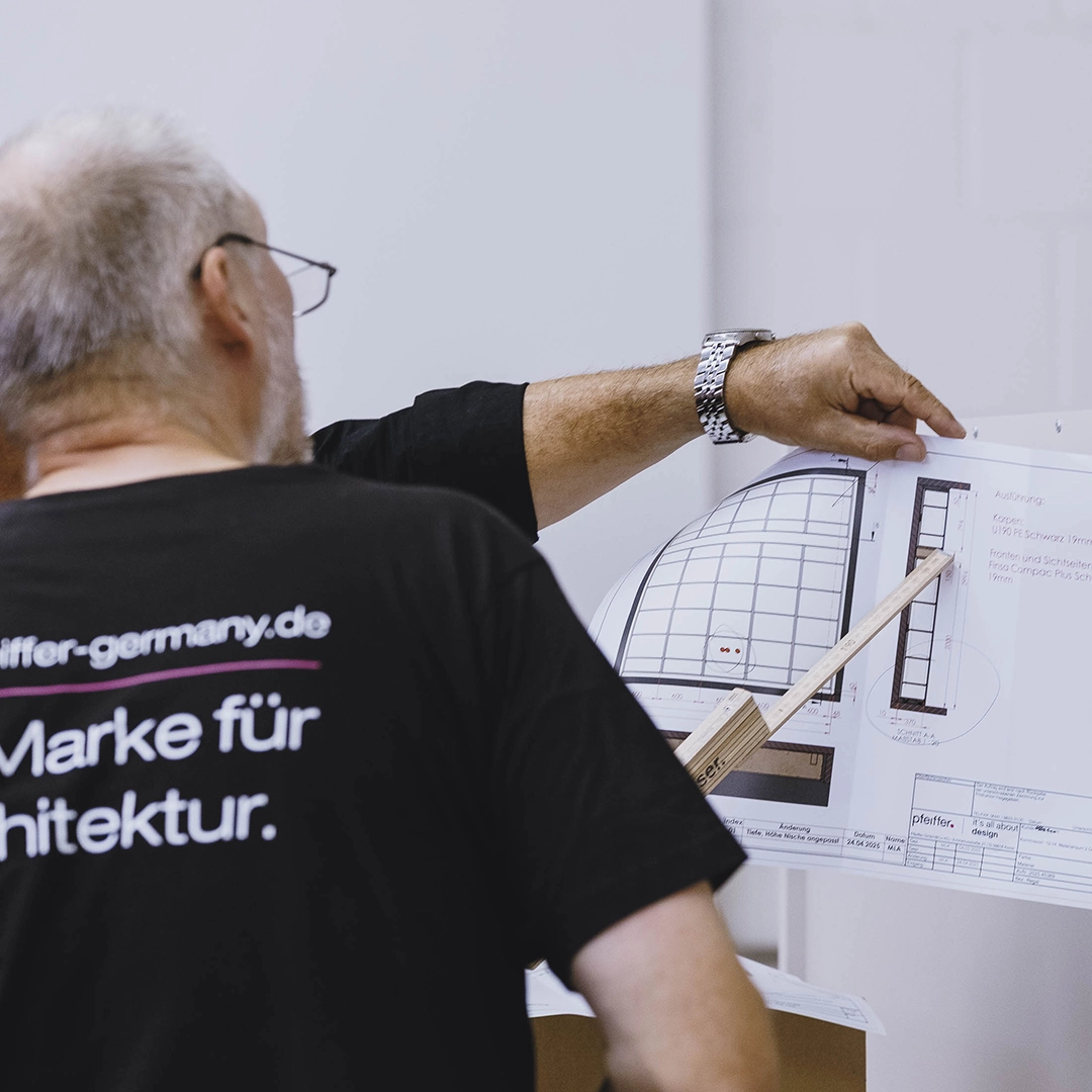 Man wearing glasses and a black shirt with text, pointing at an architectural blueprint with a wooden pointer.