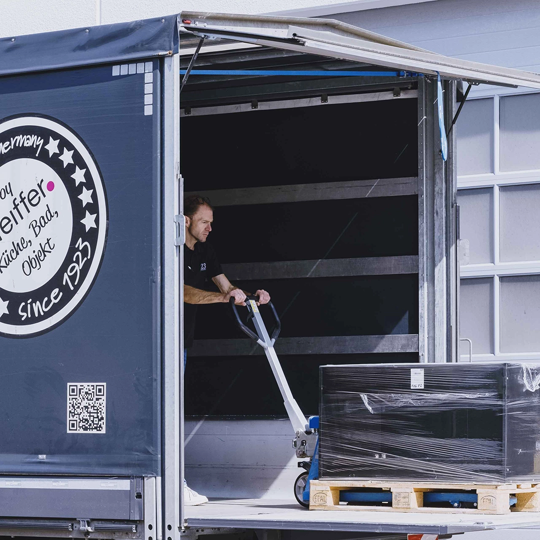 Man using a pallet jack to move a large, wrapped box on a wooden pallet inside an open delivery truck.