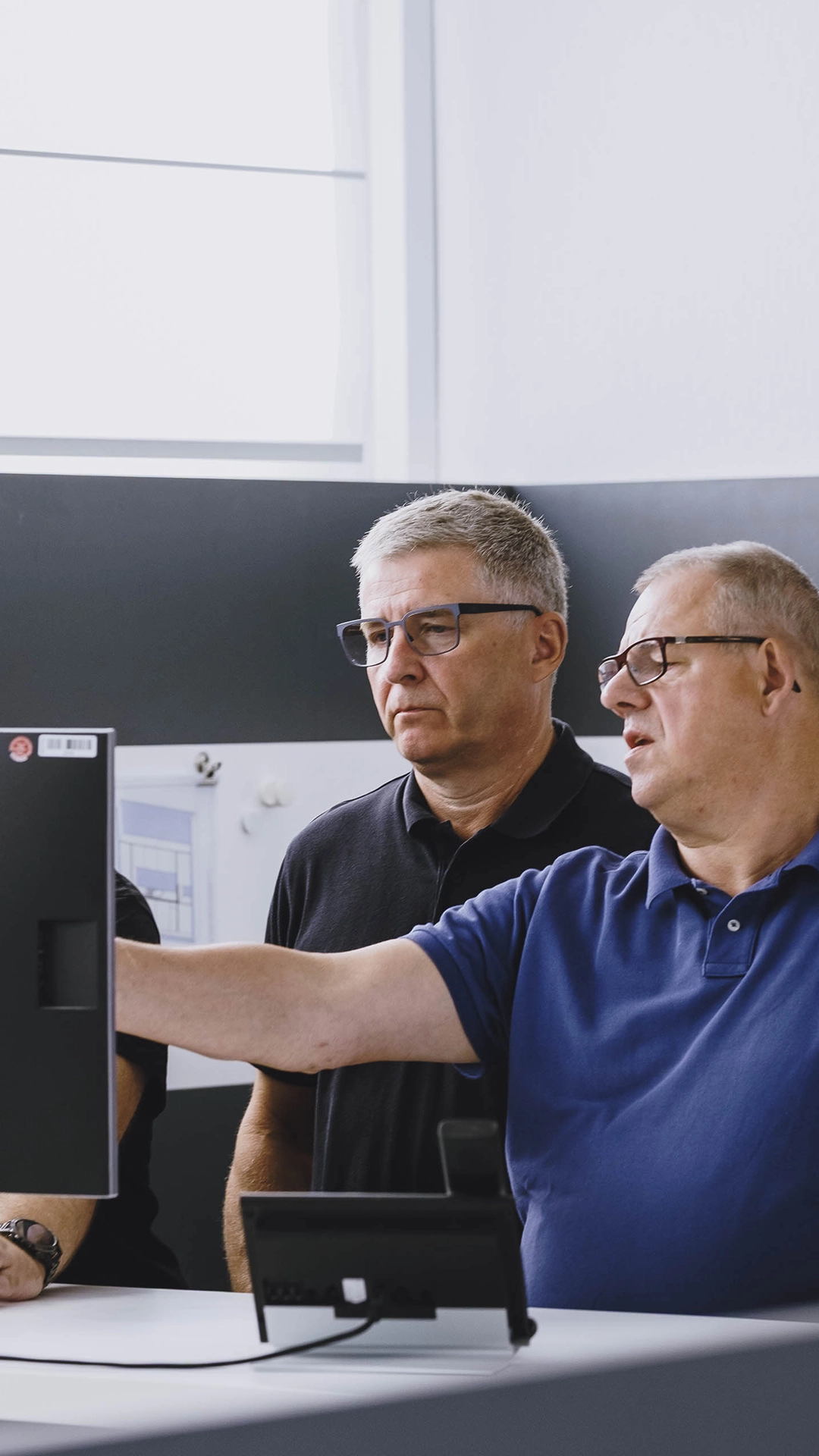 Two older men wearing glasses, one in a black shirt and one in a blue shirt, looking at a computer monitor while the man in blue points at the screen.