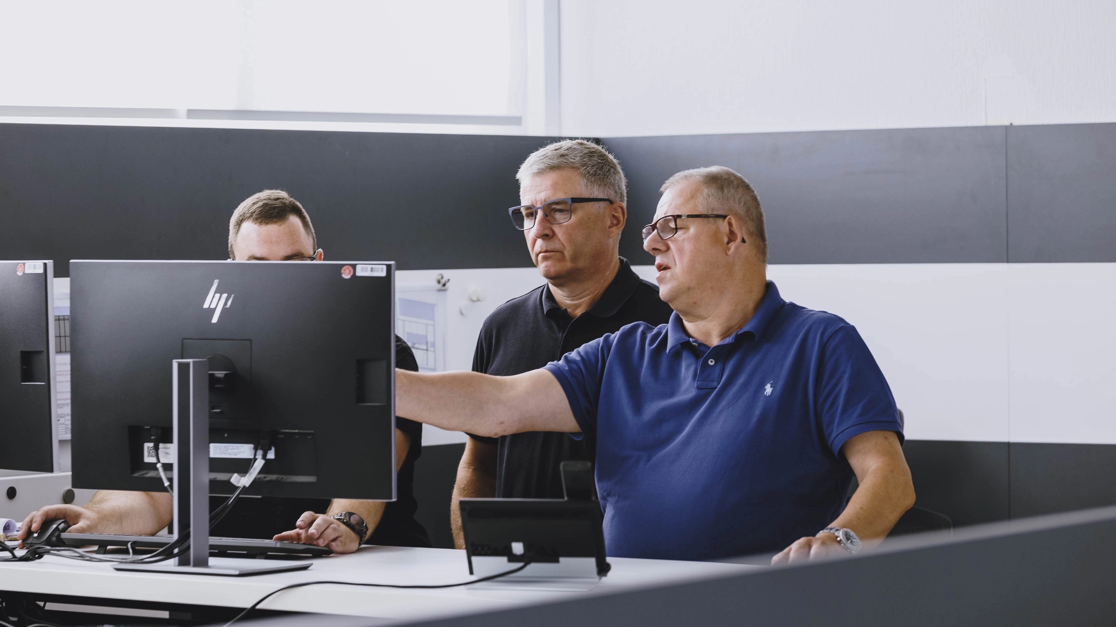 Three men working together at a desk with computer monitors, one pointing at the screen.