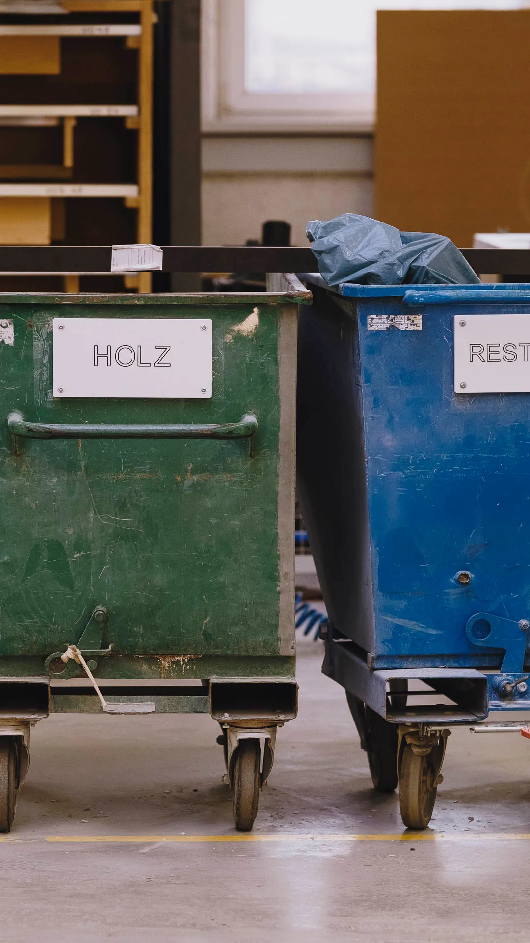 Side-by-side green and blue industrial waste bins labeled 'HOLZ' and 'REST' on wheels in a warehouse setting.