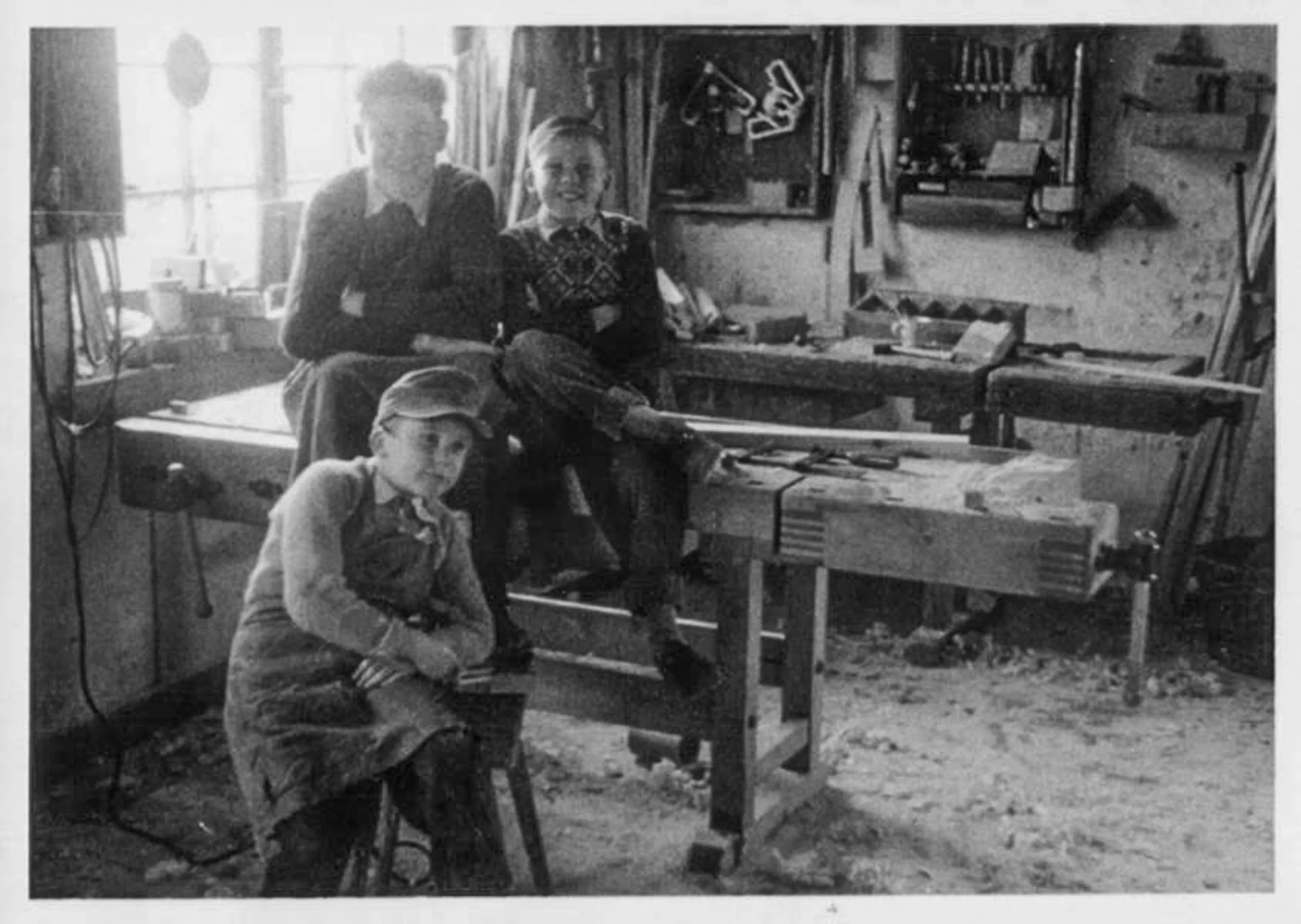Three boys in a woodworking shop, two sitting on a workbench and one seated on a stool wearing an apron and cap.