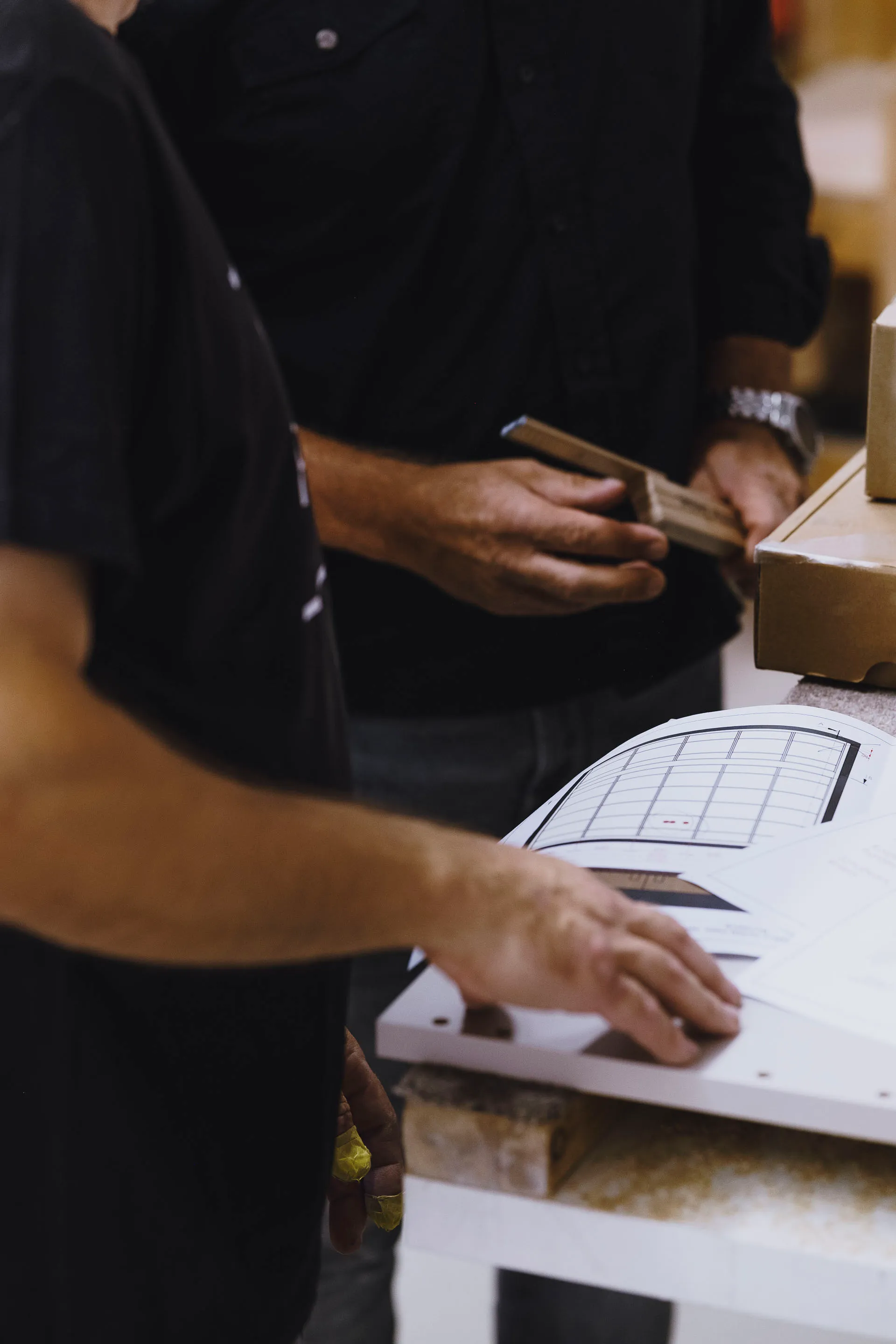 Two men examining documents and holding tools over a workbench with a cardboard box nearby.