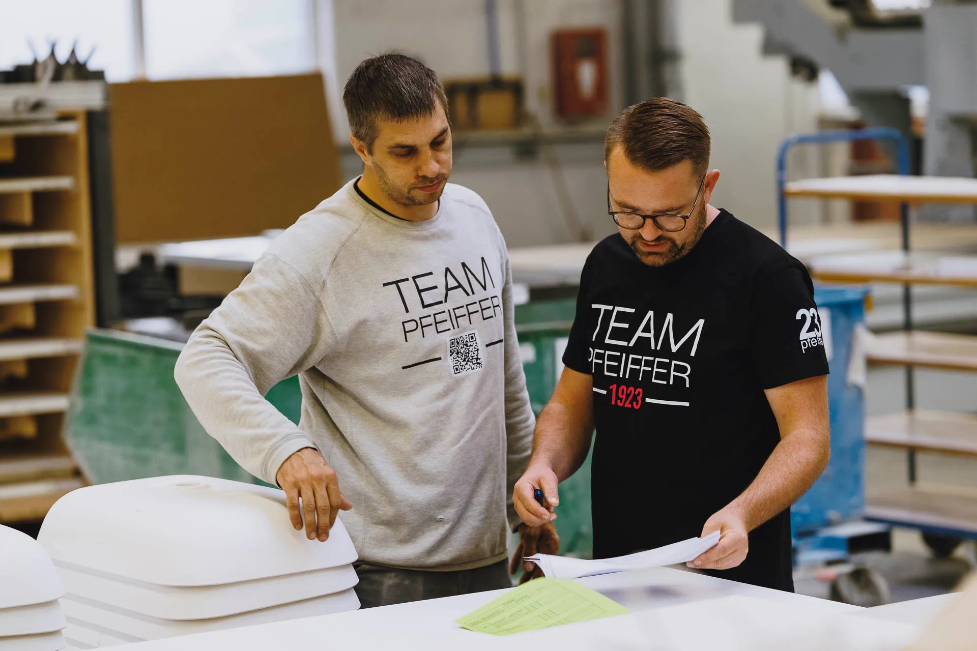 Two men in a workshop reviewing documents, both wearing 'Team Pfeiffer' shirts.