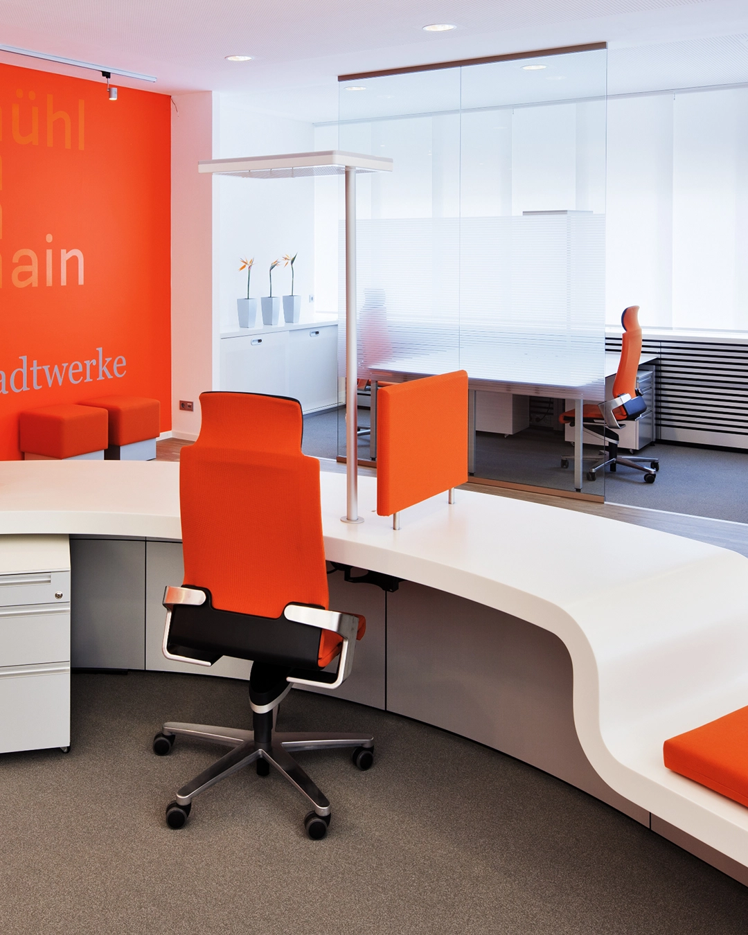 Modern office interior with a white curved desk and orange swivel chairs, orange accent wall, and glass partition.